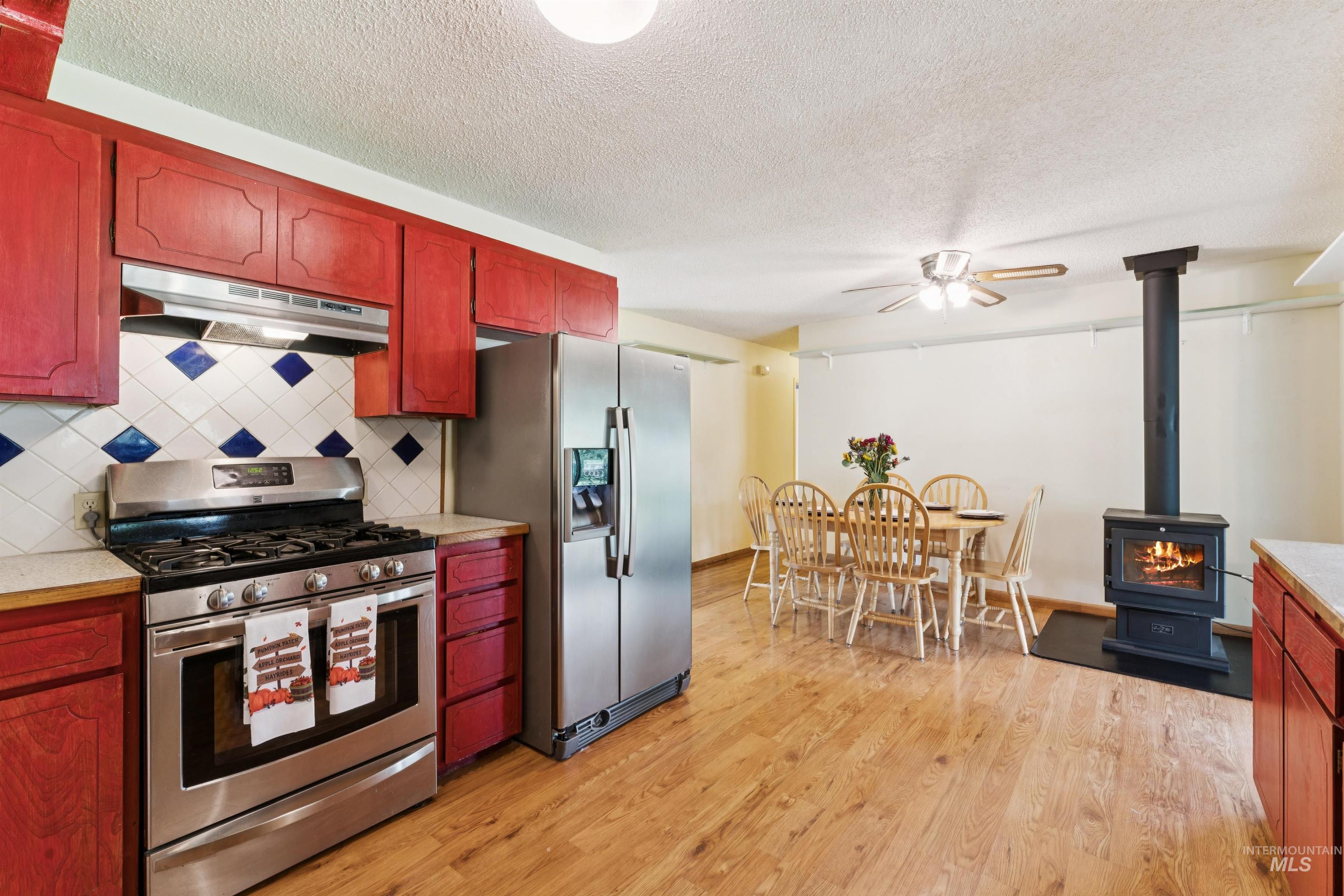 Kitchen featuring light countertops, appliances with stainless steel finishes, a wood stove, under cabinet range hood, and tasteful backsplash