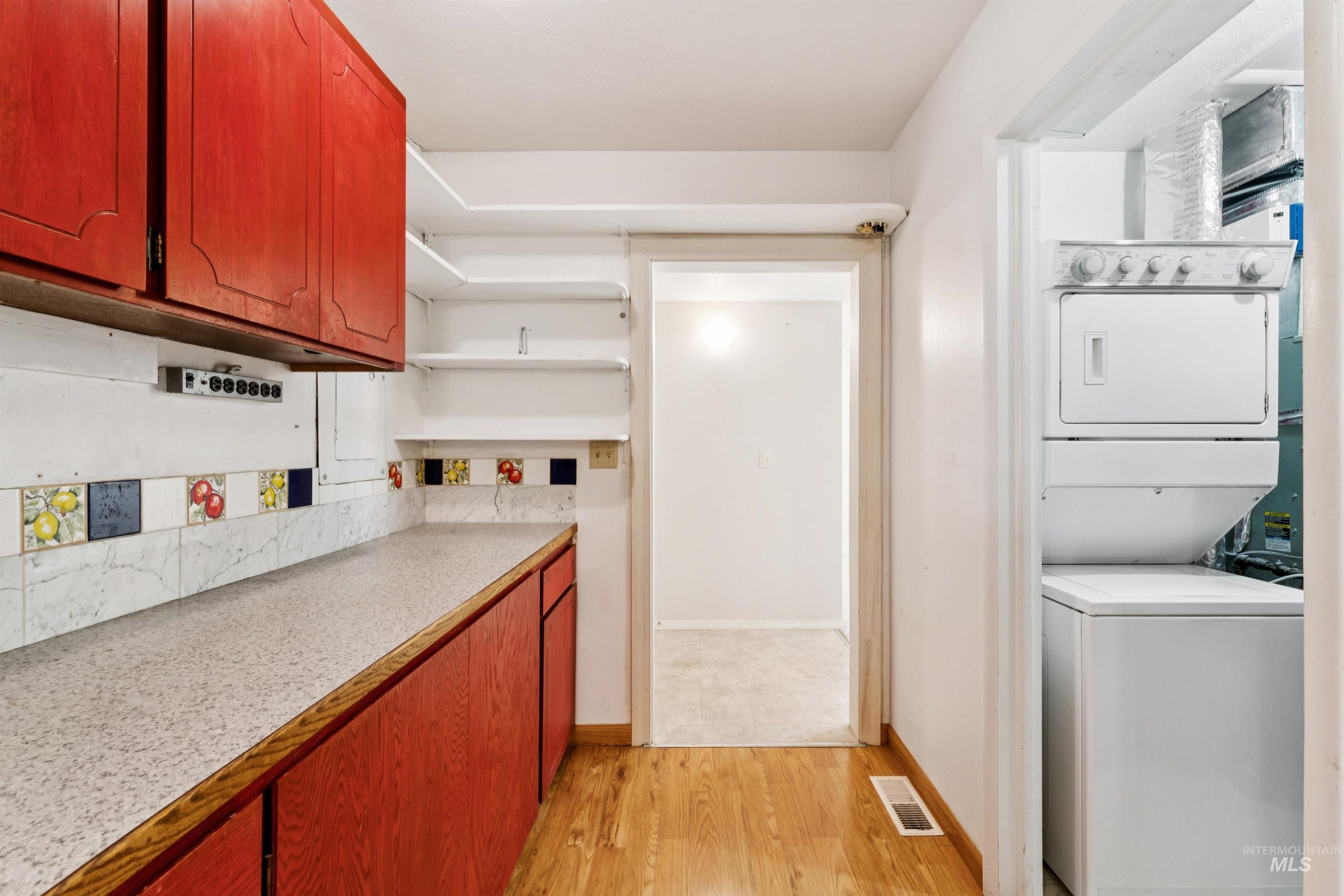 Kitchen with light countertops, light wood-type flooring, open shelves, stacked washer / drying machine, and brown cabinets