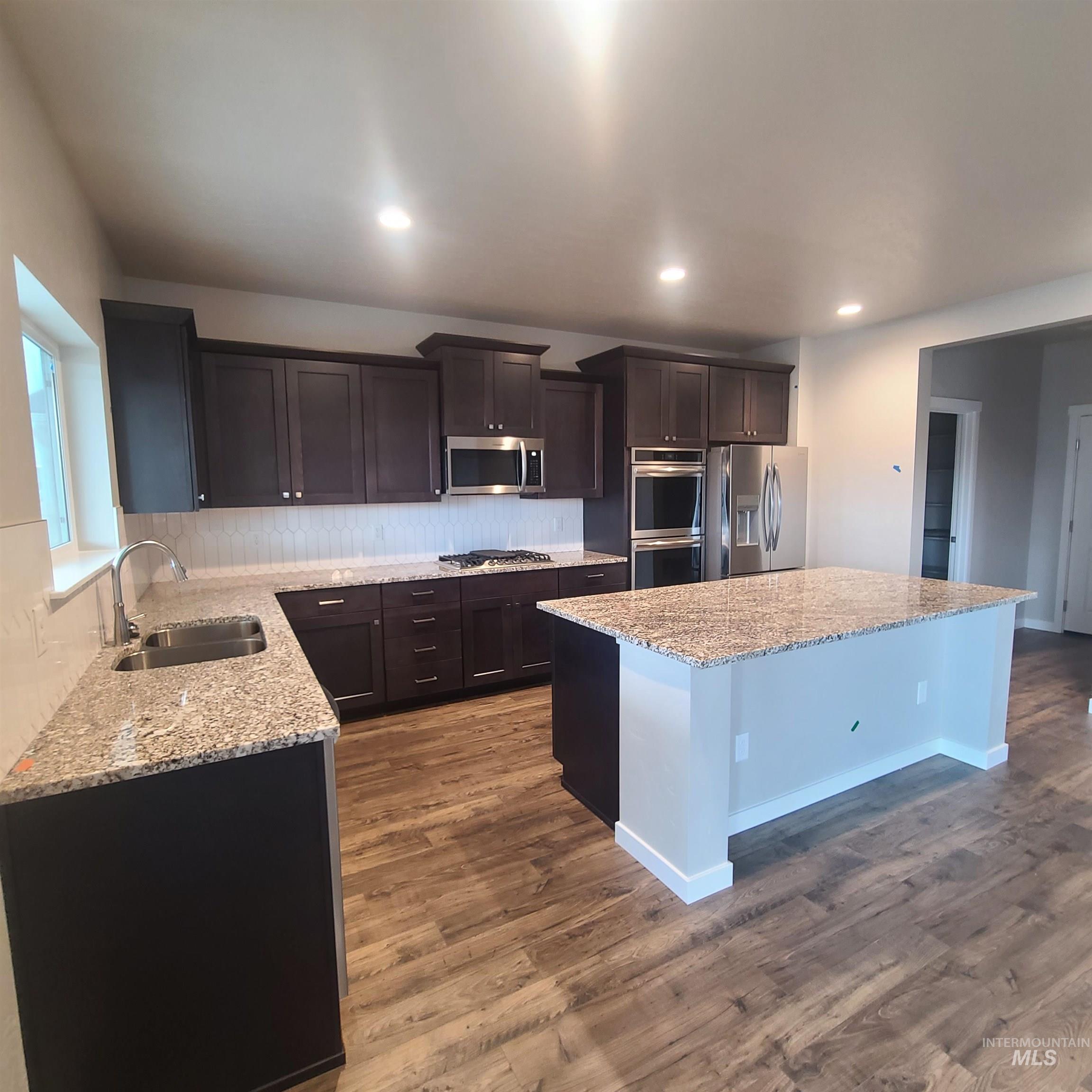 Kitchen featuring dark brown cabinets, light stone counters, decorative backsplash, stainless steel appliances, and dark wood-style floors