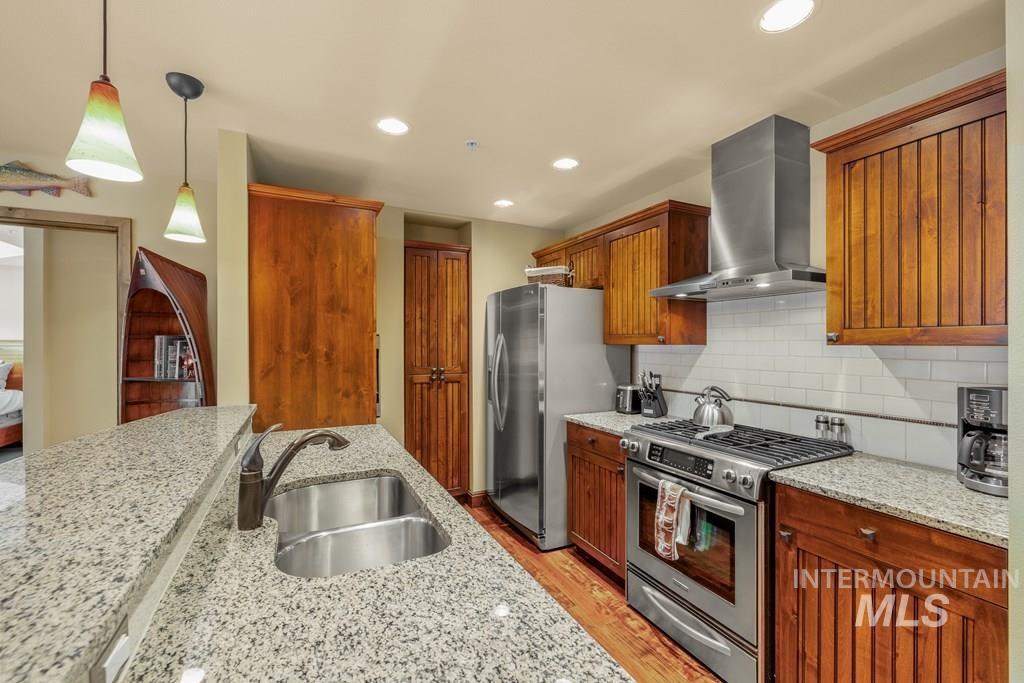 Kitchen with appliances with stainless steel finishes, wall chimney exhaust hood, pendant lighting, light stone countertops, and light wood-style floors