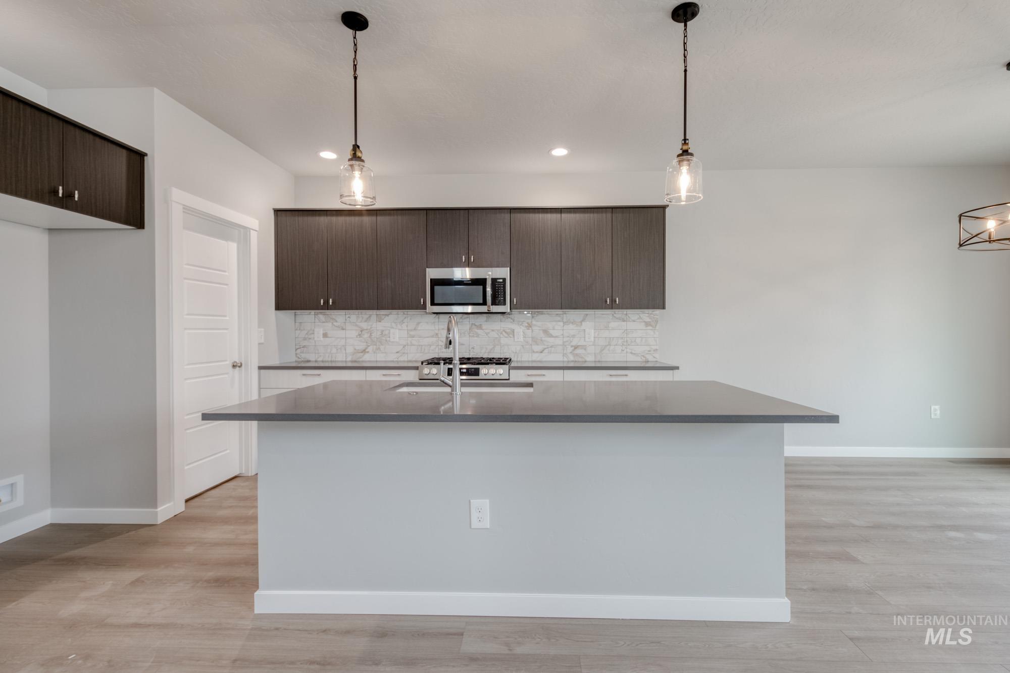 Kitchen featuring dark brown cabinetry, stainless steel microwave, tasteful backsplash, a center island with sink, and hanging light fixtures