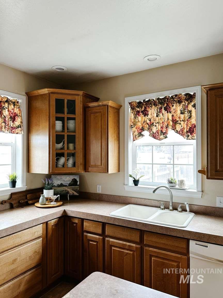 Kitchen with white dishwasher, brown cabinetry, light countertops, and glass insert cabinets