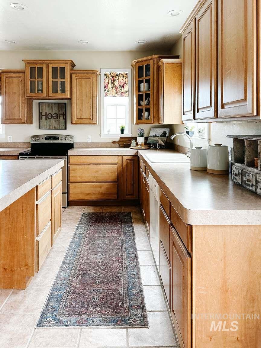 Kitchen with glass insert cabinets, stainless steel range with electric stovetop, brown cabinetry, light tile patterned floors, and light countertops