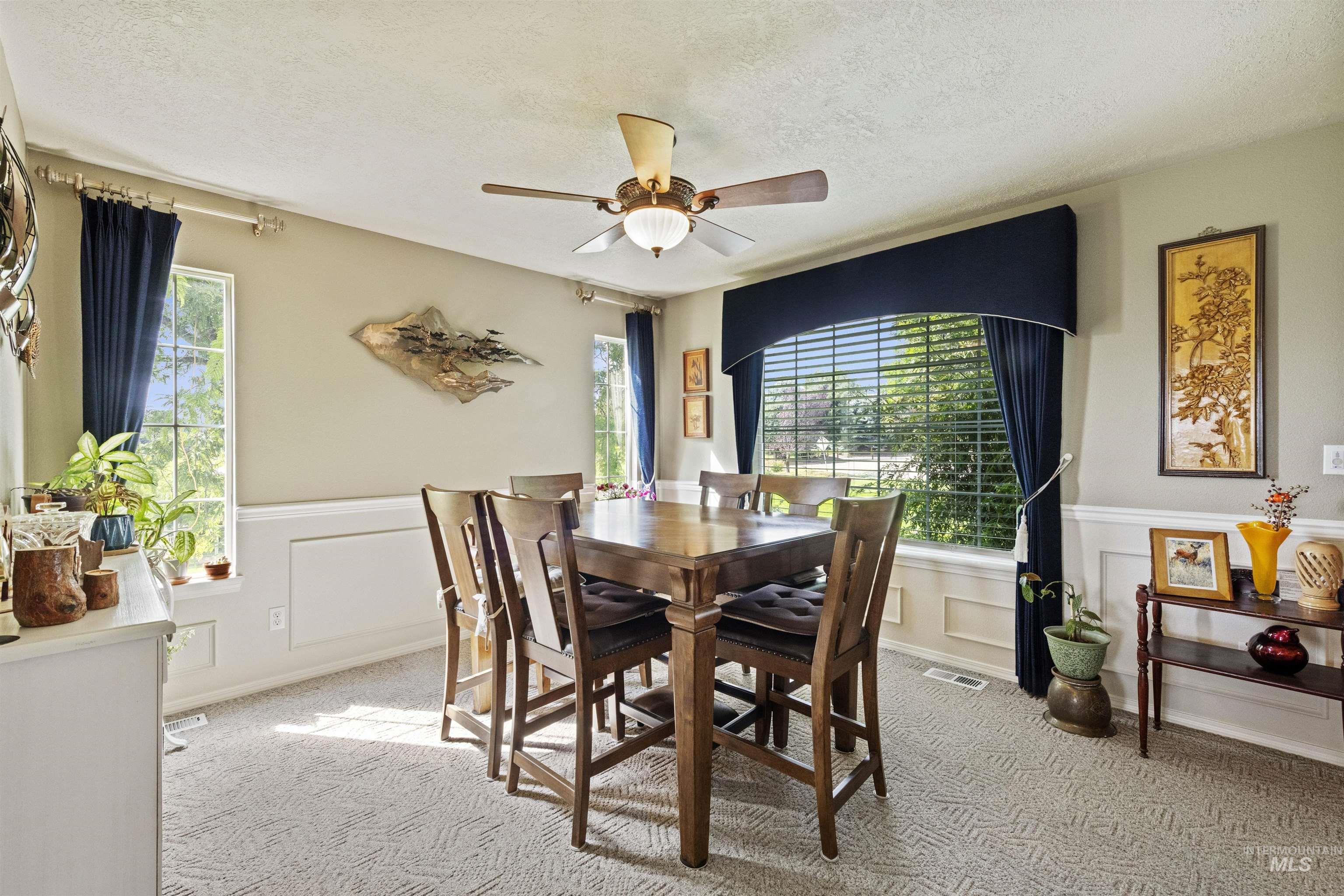 Carpeted dining area featuring healthy amount of natural light, a textured ceiling, a ceiling fan, a wainscoted wall, and a decorative wall