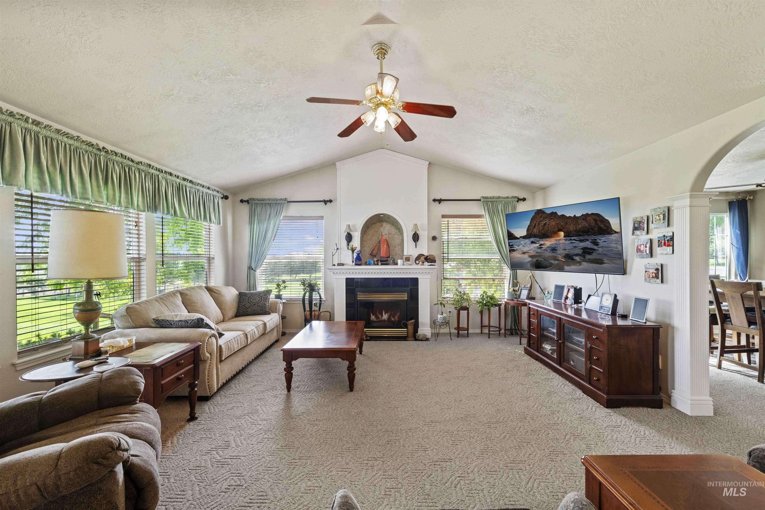 Carpeted living area with a ceiling fan, a tiled fireplace, a textured ceiling, arched walkways, and vaulted ceiling