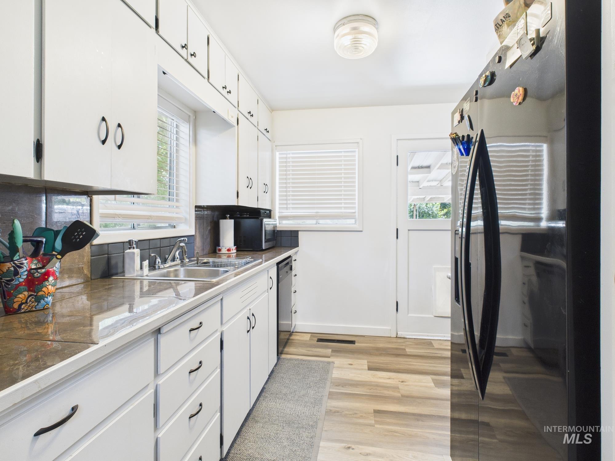 Kitchen featuring black refrigerator with ice dispenser, white cabinetry, light wood-type flooring, dishwasher, and decorative backsplash