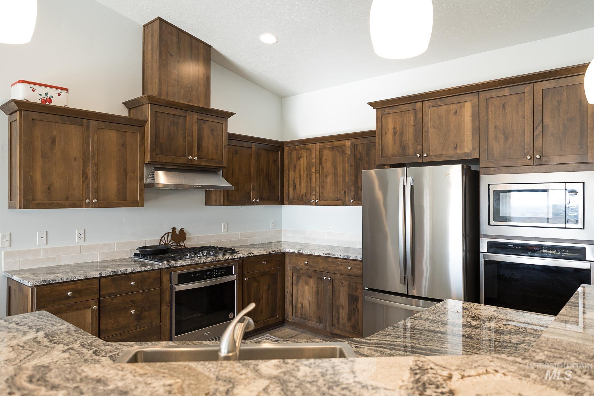 Kitchen featuring stainless steel appliances, light stone countertops, under cabinet range hood, vaulted ceiling, and dark brown cabinetry