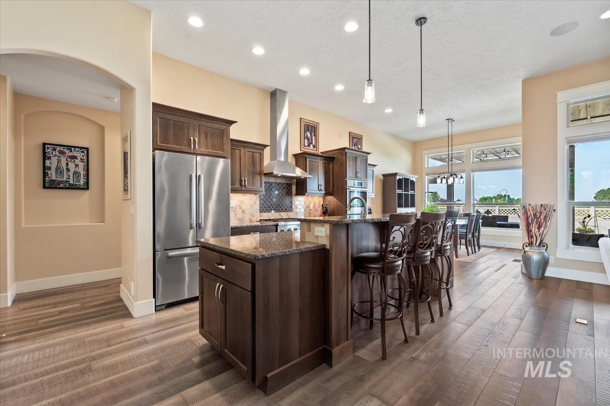 Kitchen featuring dark stone countertops, stainless steel fridge, a kitchen bar, hanging light fixtures, and wall chimney range hood