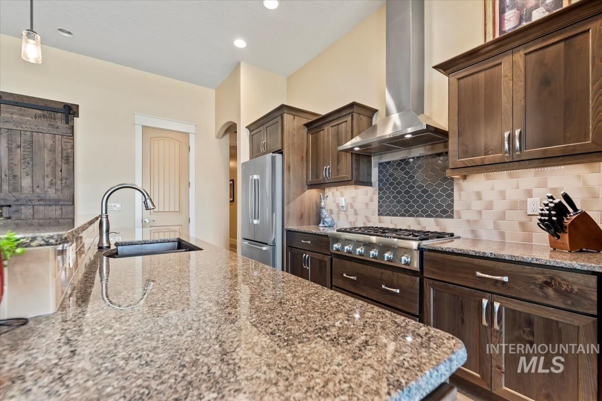 Kitchen with dark brown cabinets, wall chimney exhaust hood, tasteful backsplash, light stone counters, and stainless steel appliances