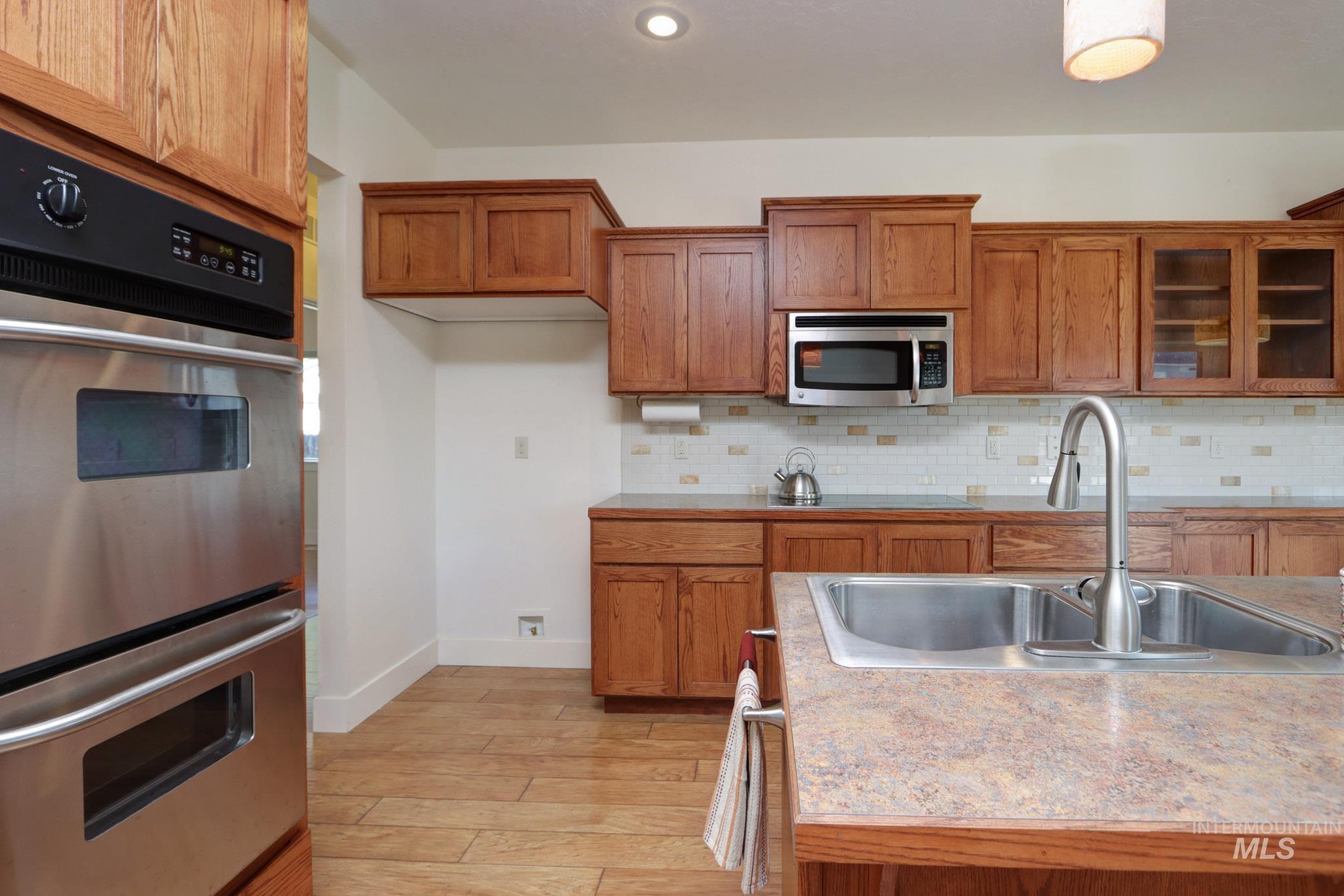 Kitchen featuring appliances with stainless steel finishes, brown cabinets, glass insert cabinets, light wood-type flooring, and backsplash