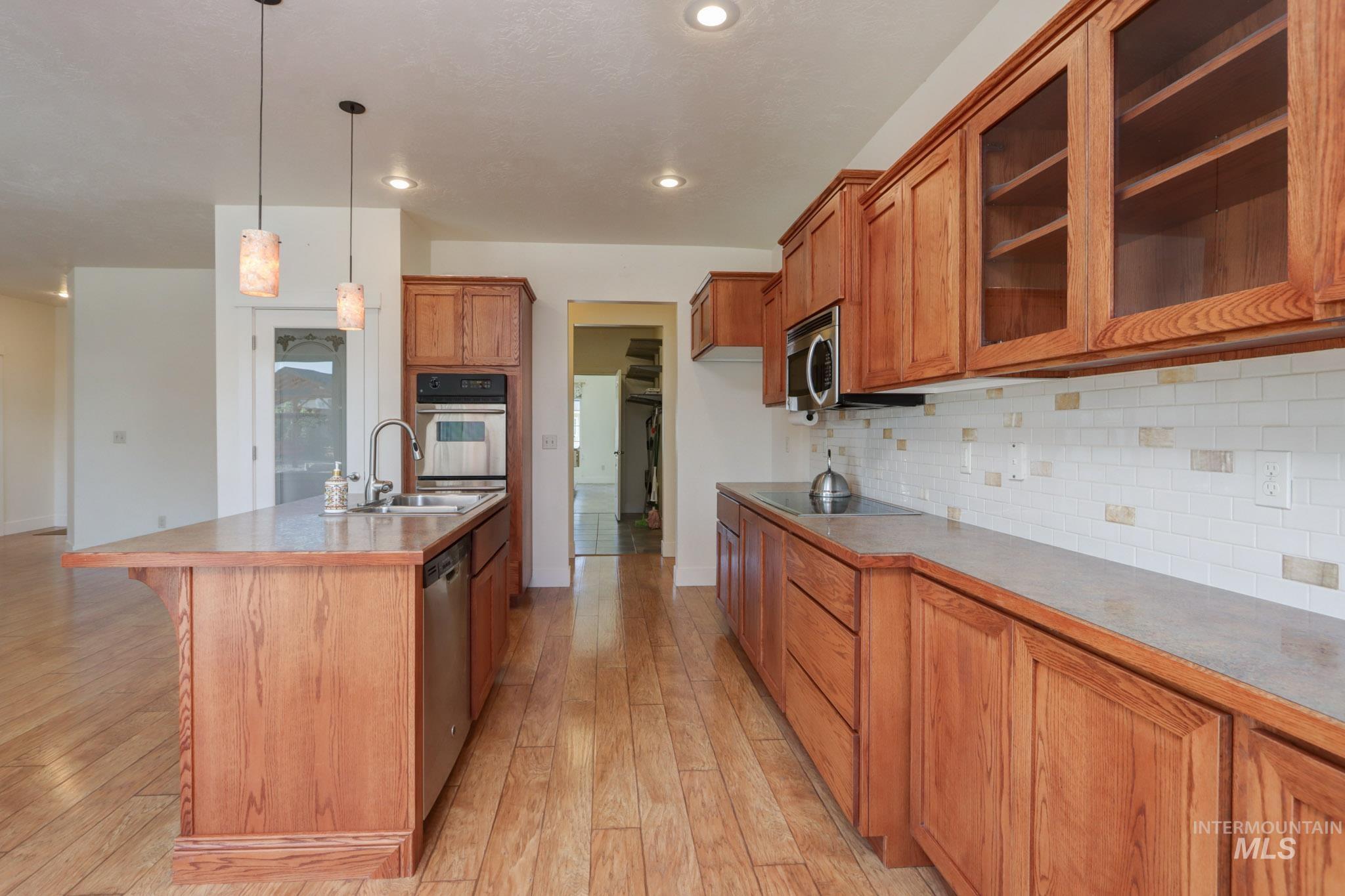 Kitchen with brown cabinetry, appliances with stainless steel finishes, a center island with sink, glass insert cabinets, and hanging light fixtures