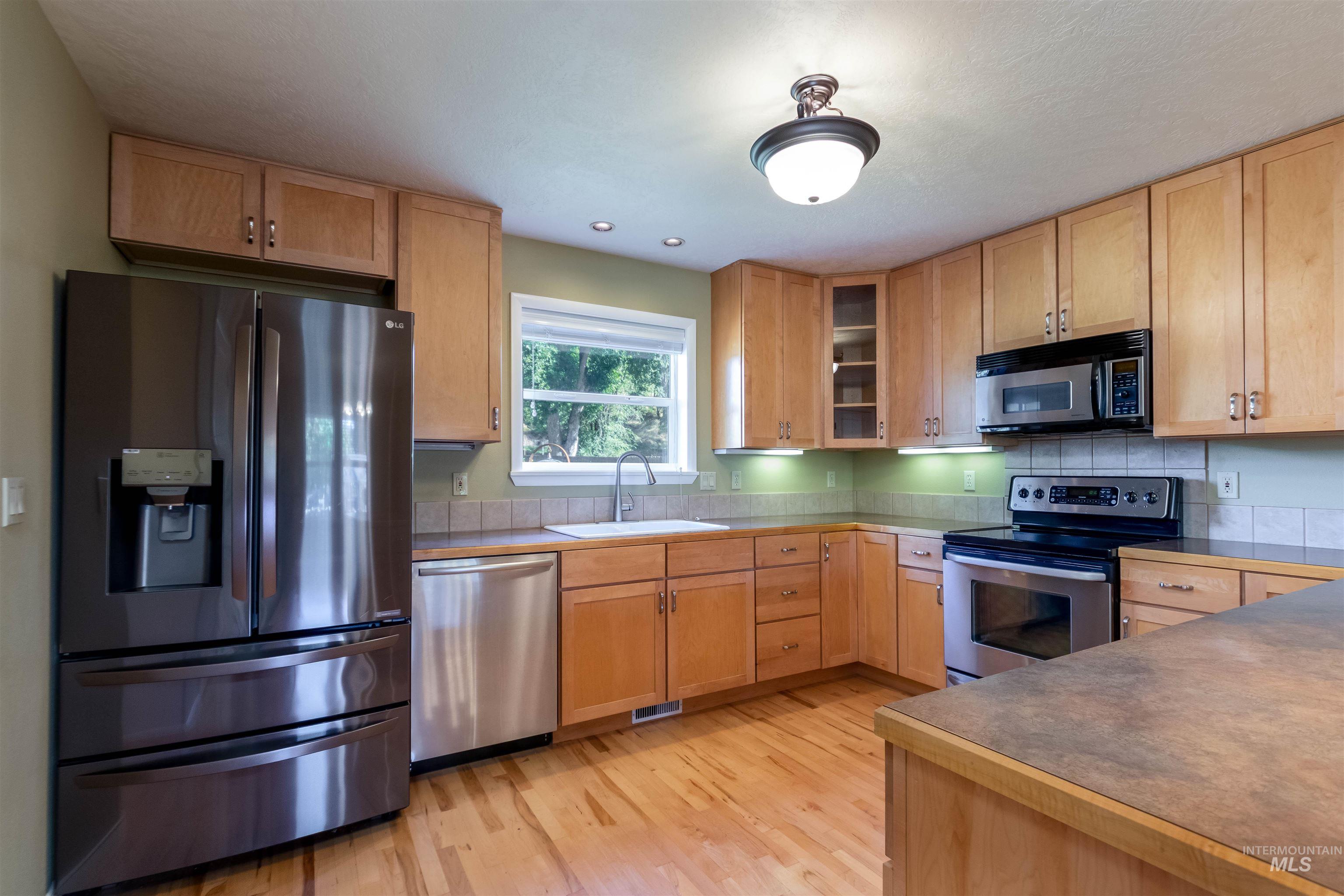 Kitchen with stainless steel appliances, light maple flooring, dark countertops, and glass insert cabinets
