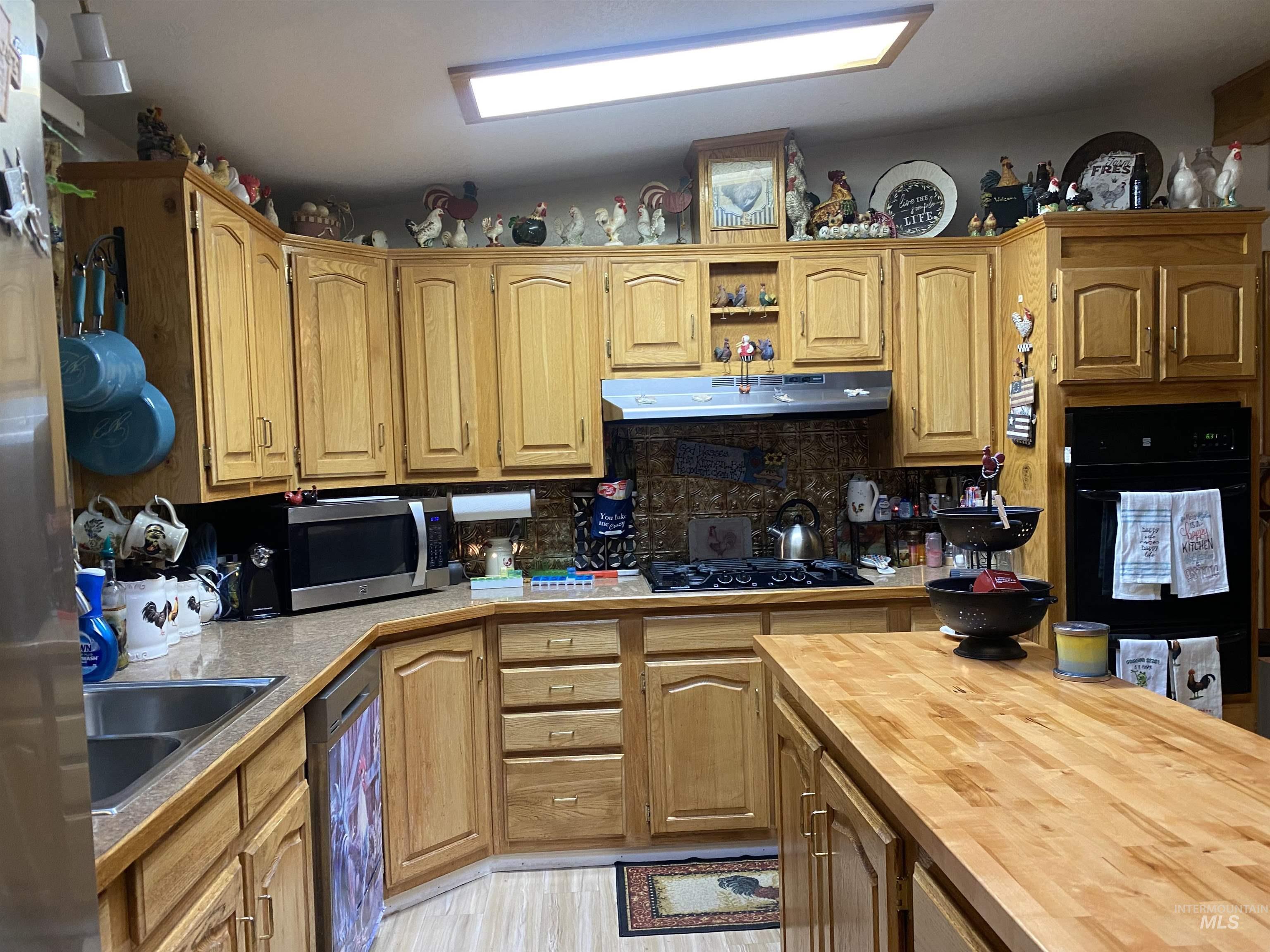 Kitchen with black appliances, backsplash, under cabinet range hood, open shelves, and brown cabinetry
