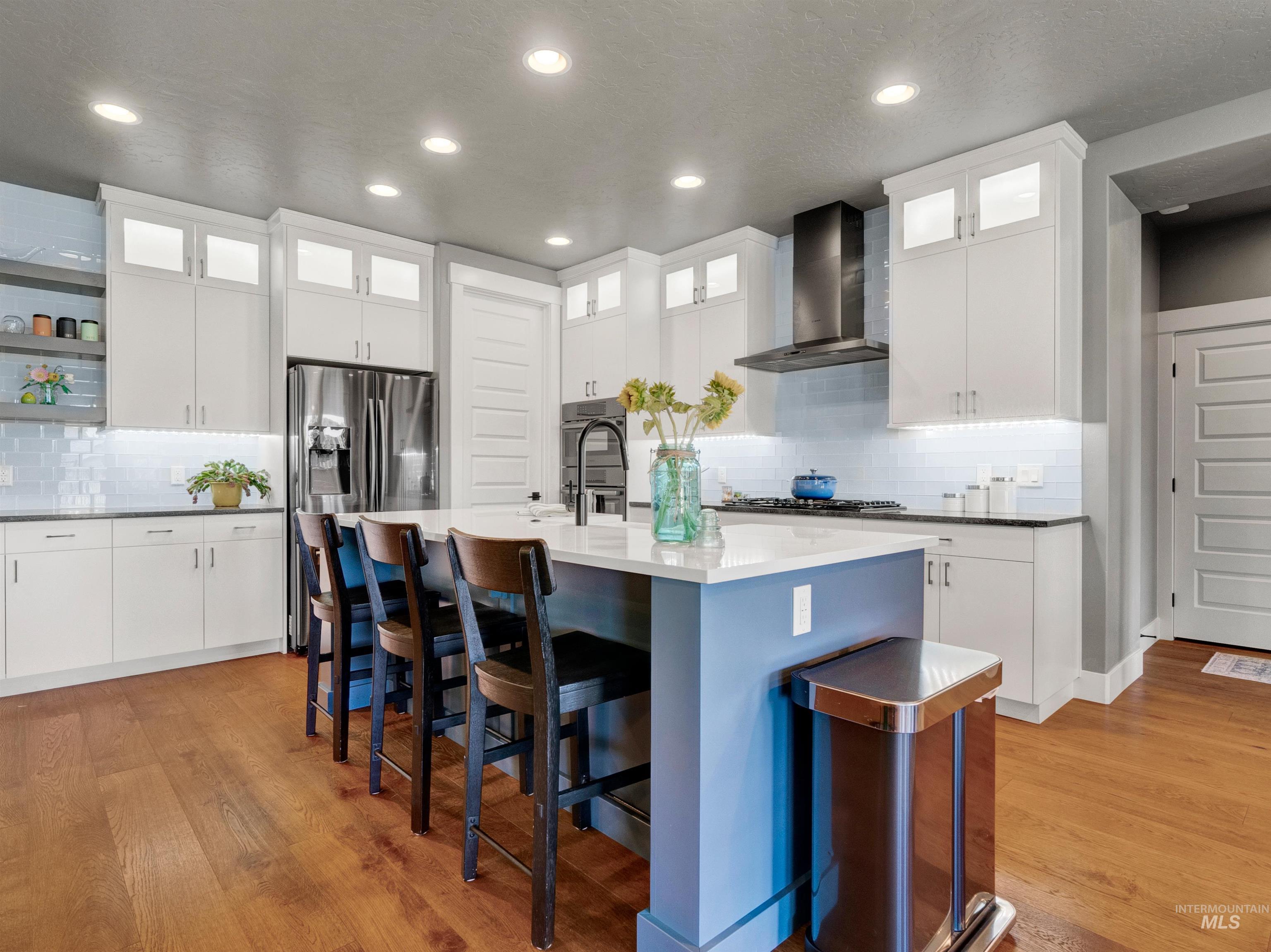 Kitchen featuring decorative backsplash, glass insert cabinets, white cabinetry, wall chimney range hood, and light wood-style flooring