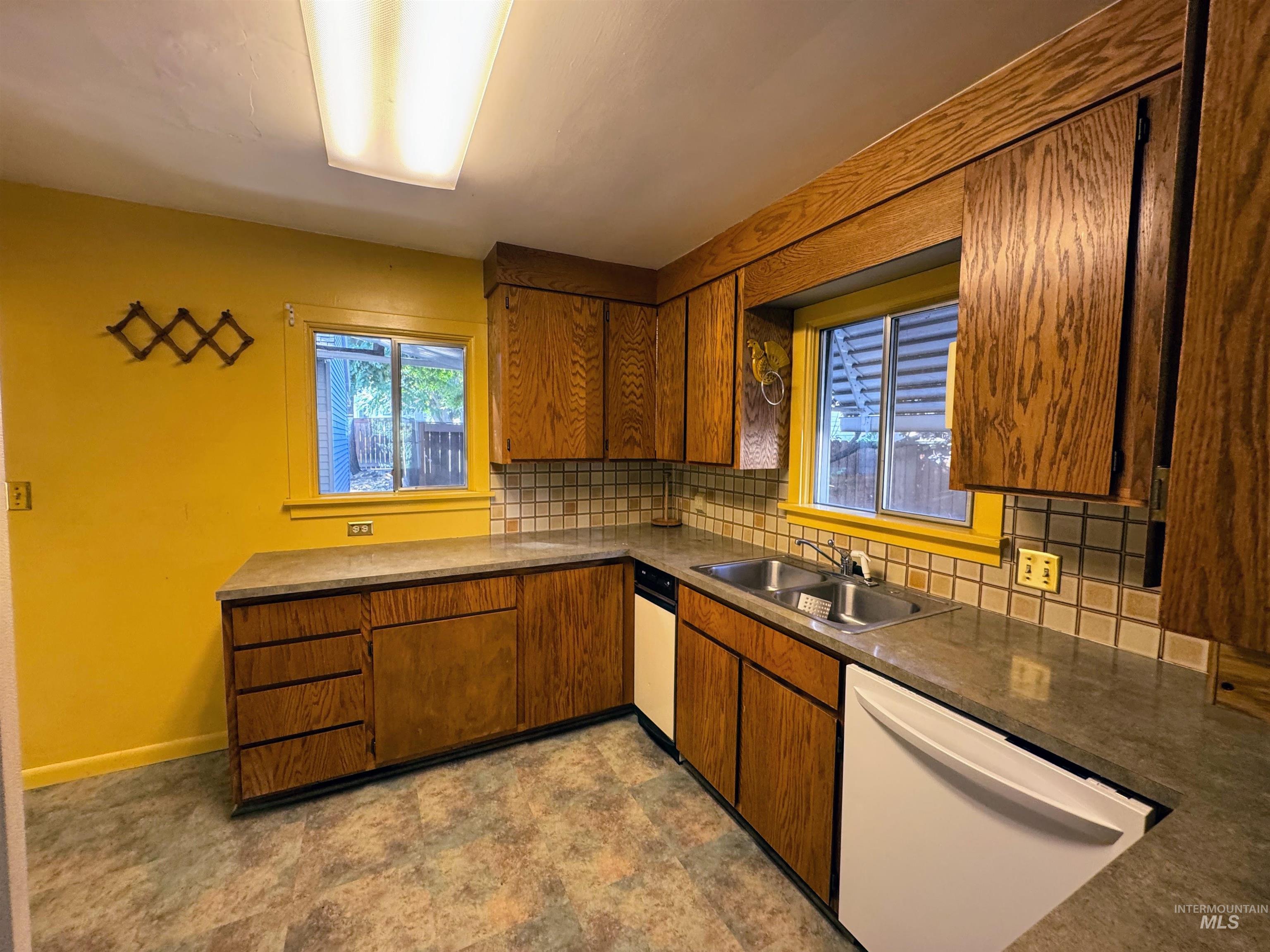 Kitchen with decorative backsplash, dishwasher, brown cabinets, dark countertops, and stone finish floors