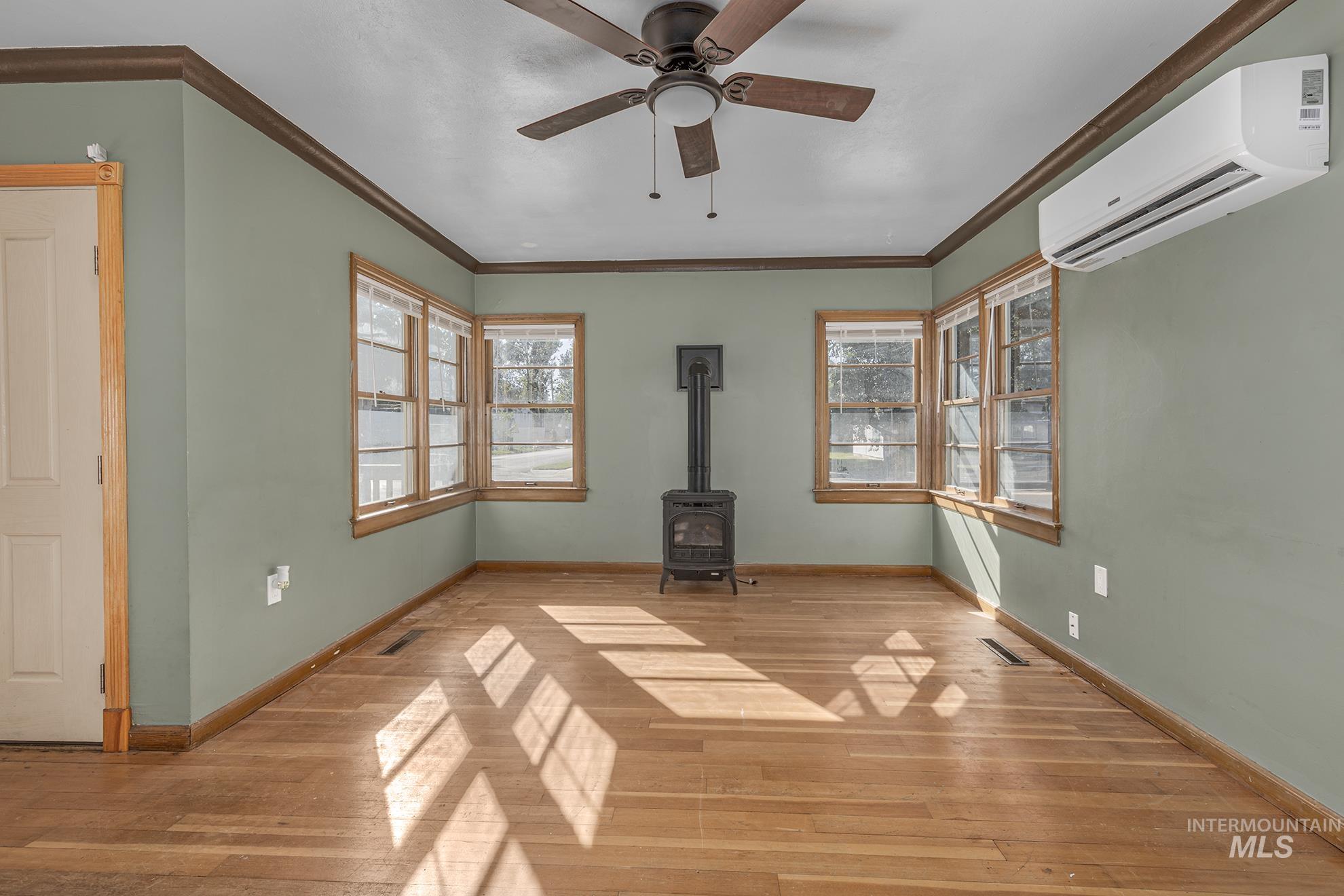 Unfurnished living room with a wood stove, plenty of natural light, a wall mounted AC, ornamental molding, and hardwood / wood-style floors