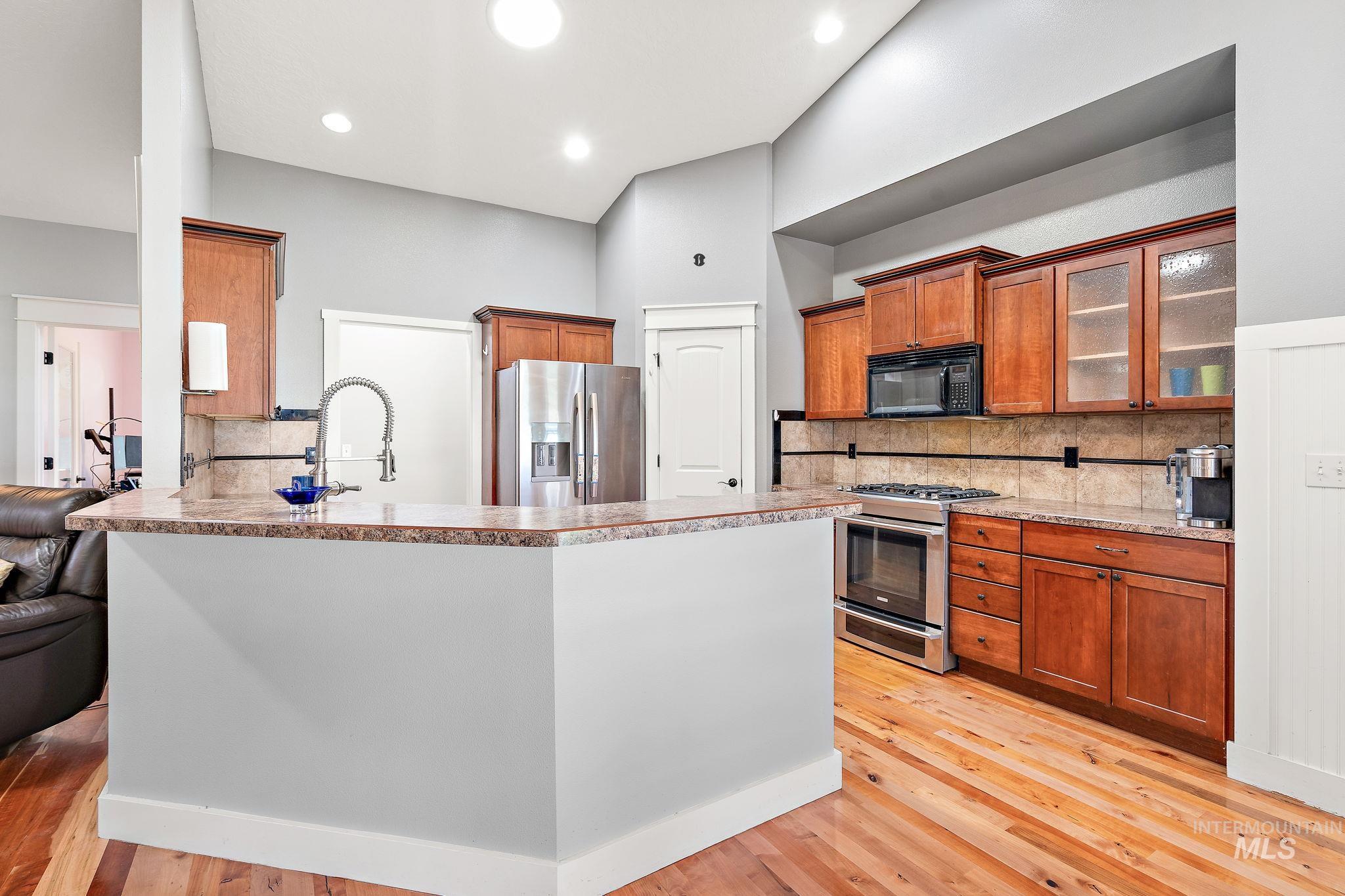 Kitchen with brown cabinets, decorative backsplash, appliances with stainless steel finishes, light wood-type flooring, and glass insert cabinets