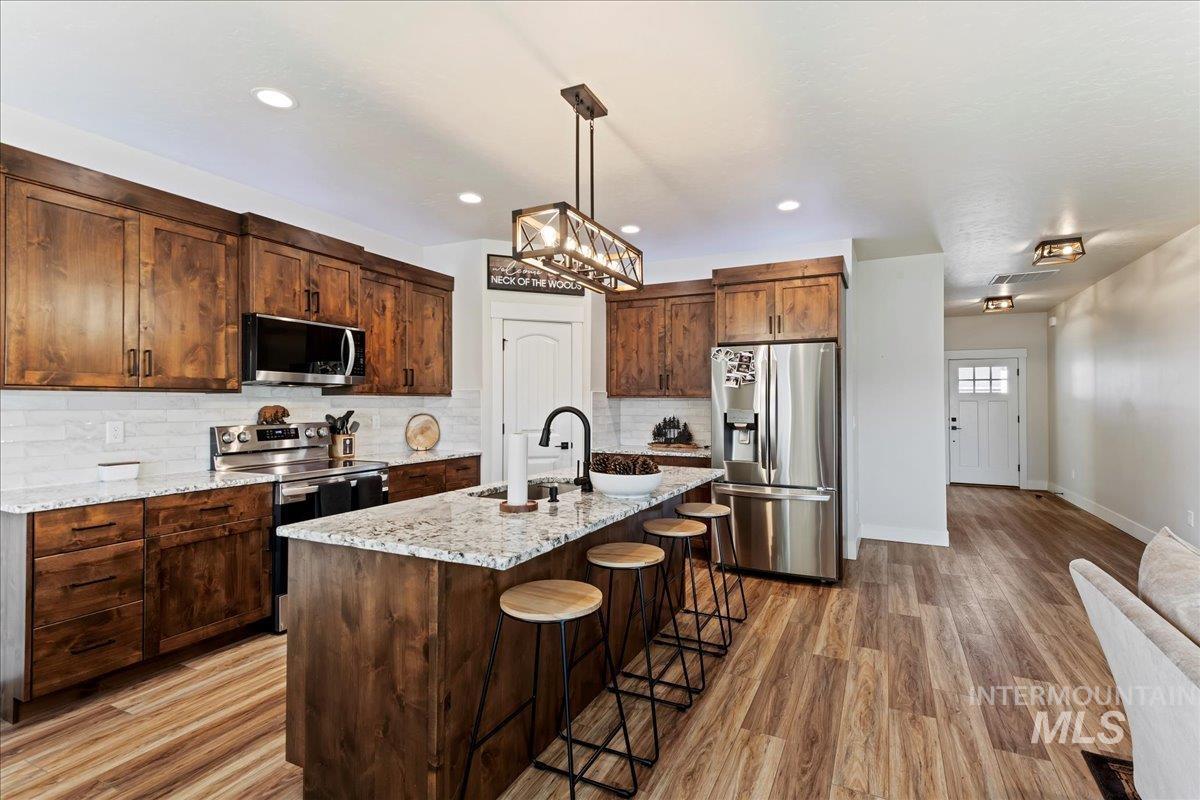 Kitchen with appliances with stainless steel finishes, backsplash, decorative light fixtures, light wood finished floors, and a kitchen island with sink