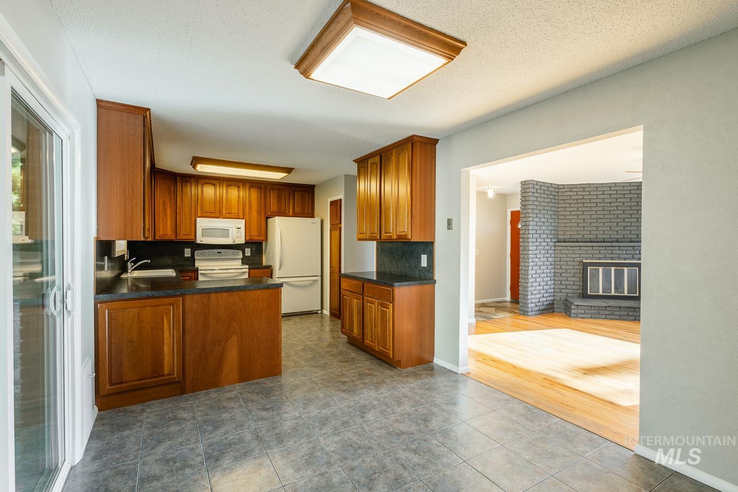 Kitchen featuring brown cabinets, white appliances, a peninsula, a brick fireplace, and dark tile patterned floors