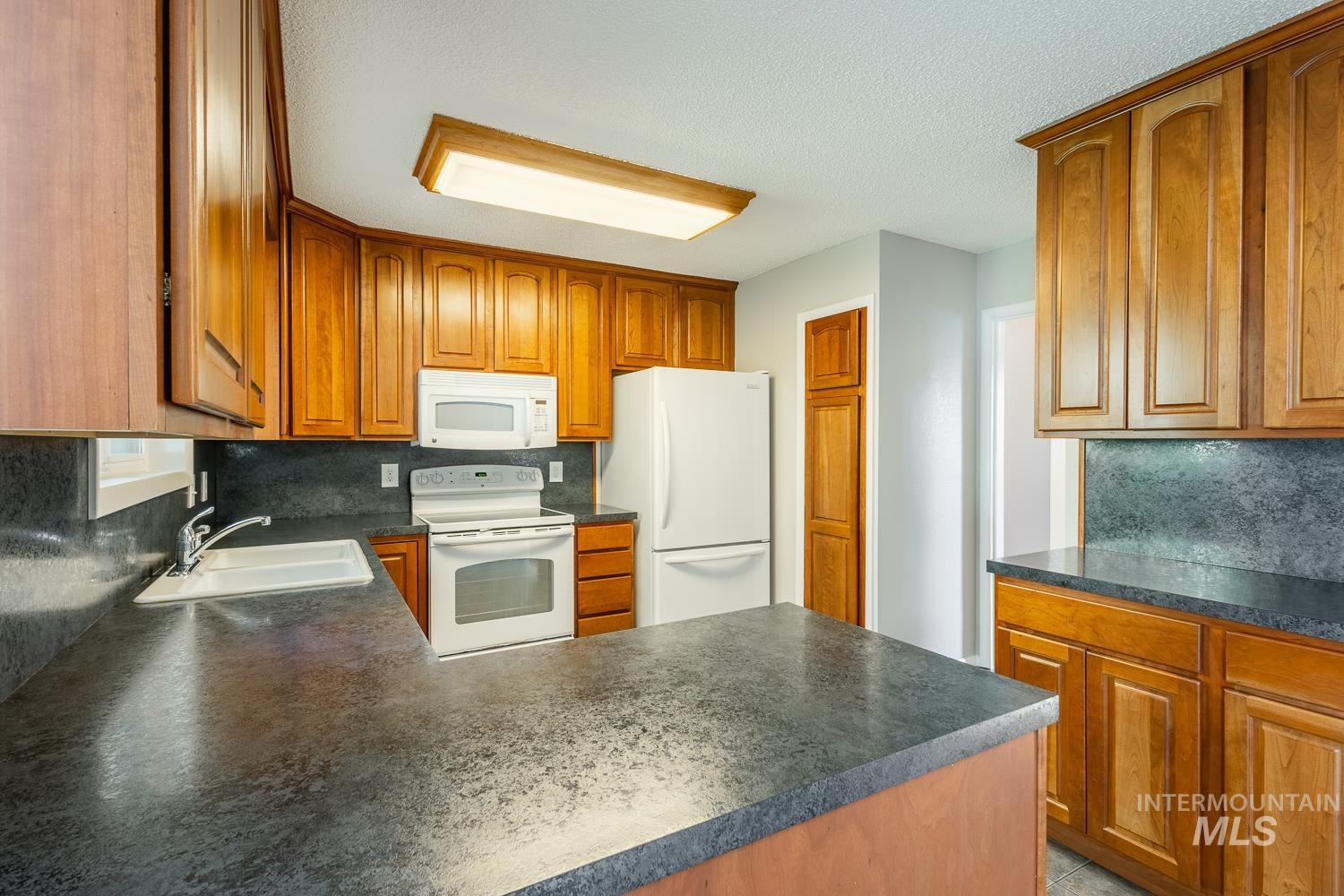 Kitchen with dark countertops, a peninsula, backsplash, white appliances, and brown cabinetry