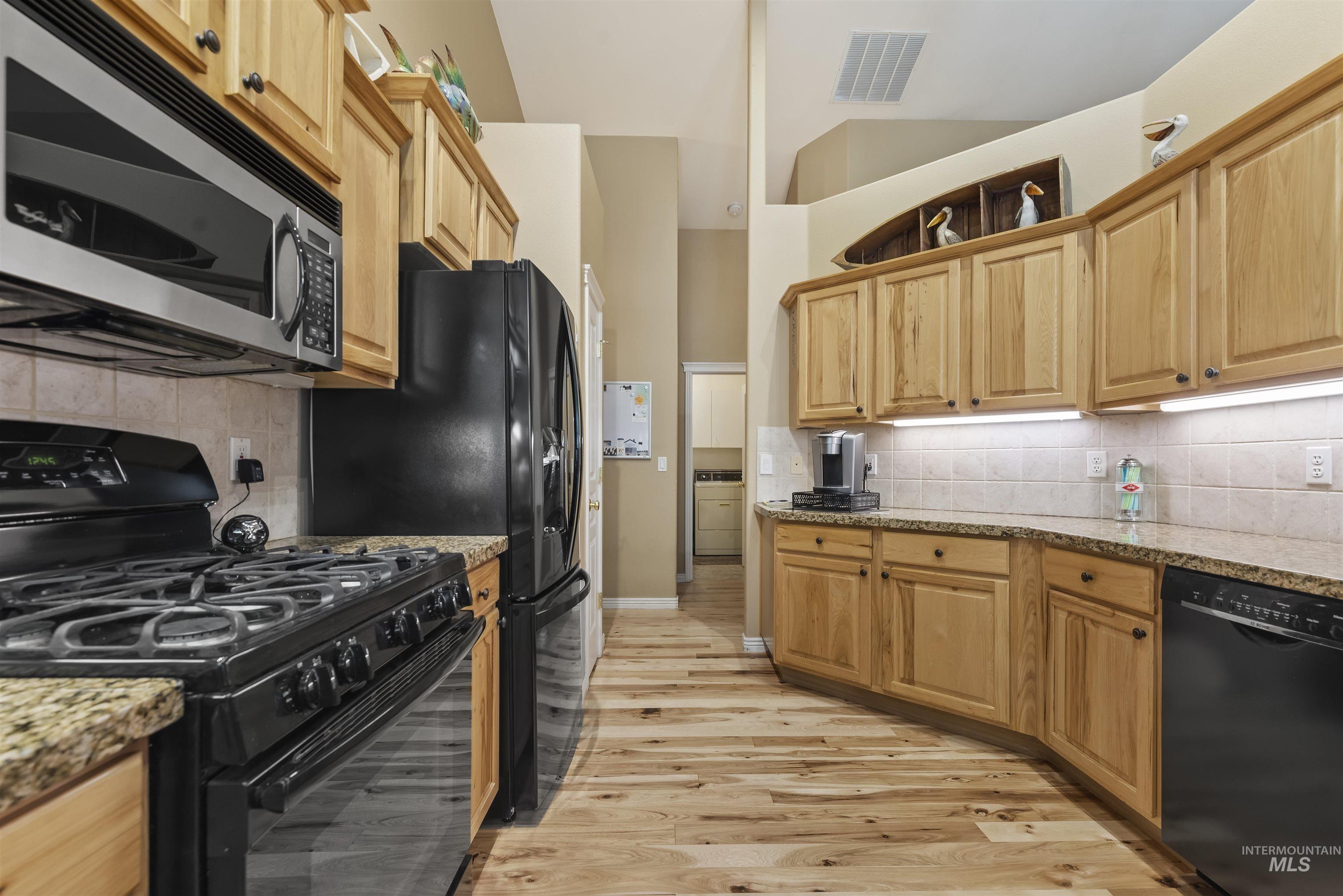 Kitchen with black appliances, tasteful backsplash, light stone counters, light wood-style flooring, and a towering ceiling