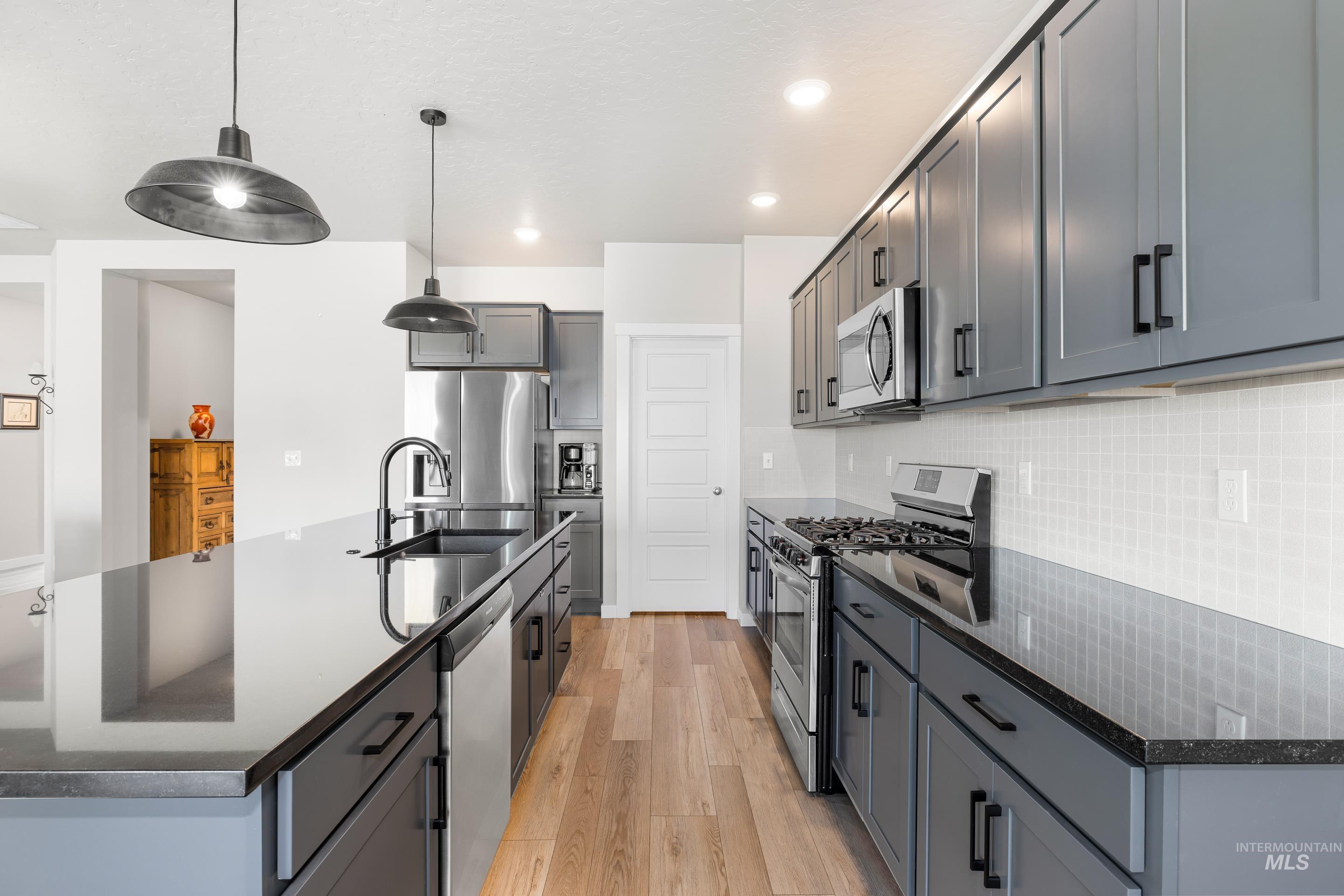 Kitchen featuring stainless steel appliances, a kitchen island with sink, light wood finished floors, decorative light fixtures, and dark stone counters