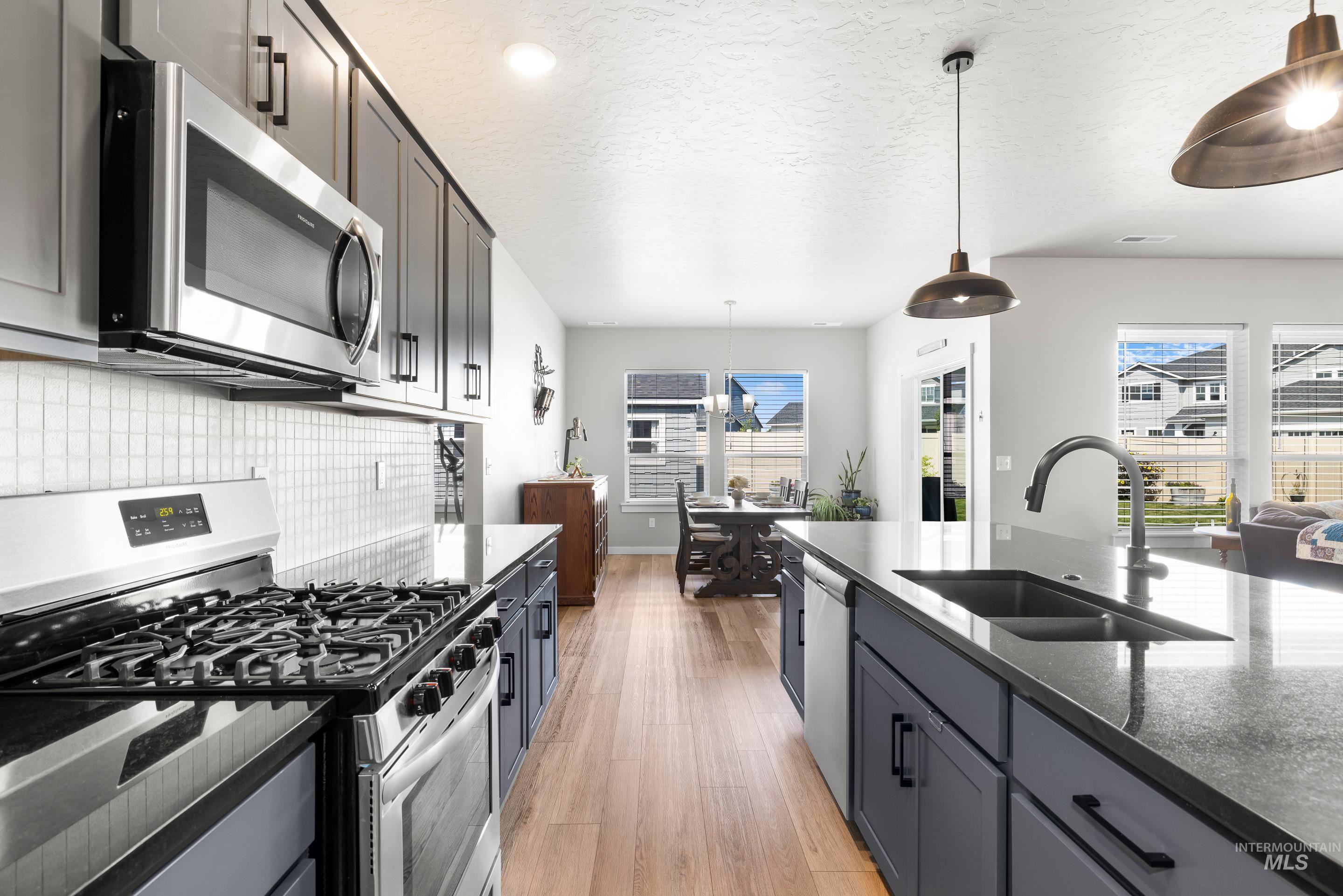 Kitchen featuring stainless steel appliances, hanging light fixtures, light wood-style floors, dark stone counters, and plenty of natural light