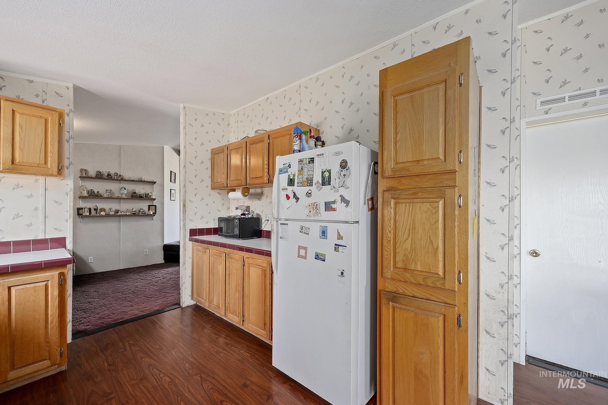 Kitchen with tile countertops, freestanding refrigerator, dark wood finished floors, wallpapered walls, and black microwave