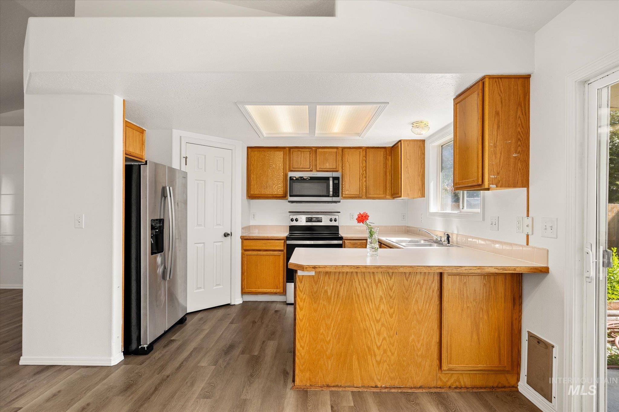 Kitchen with a peninsula, light countertops, dark wood-type flooring, appliances with stainless steel finishes, and brown cabinets