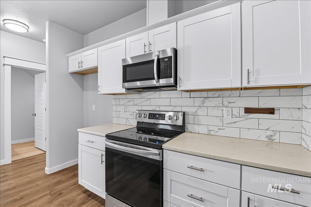 Kitchen with stainless steel appliances, tasteful backsplash, white cabinetry, light wood-type flooring, and light stone counters