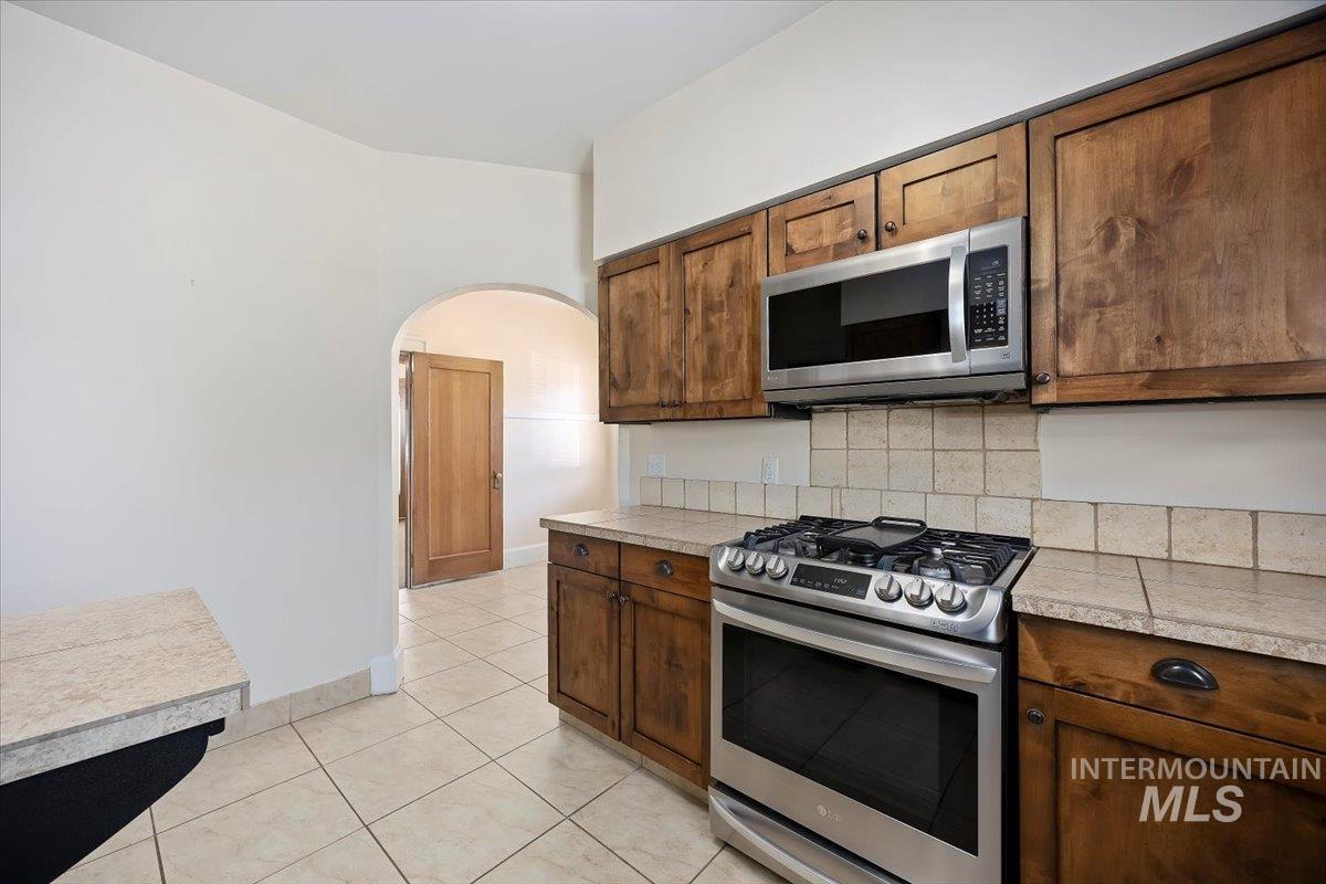 Kitchen with appliances with stainless steel finishes, light tile patterned flooring, arched walkways, backsplash, and brown cabinetry