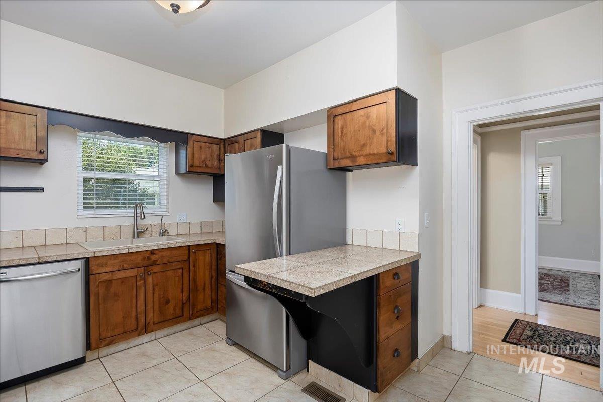 Kitchen featuring stainless steel appliances, light tile patterned floors, tile counters, and brown cabinets