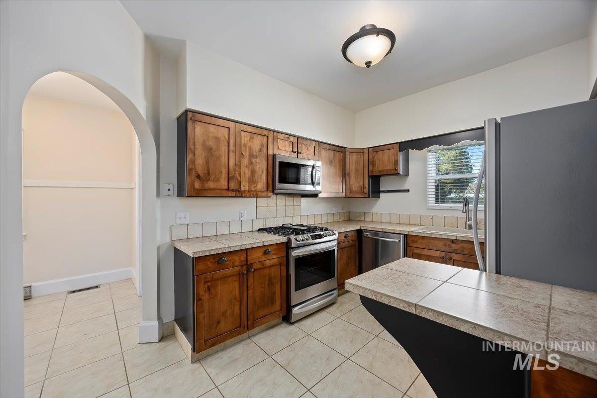 Kitchen with stainless steel appliances, brown cabinets, light tile patterned floors, and arched walkways