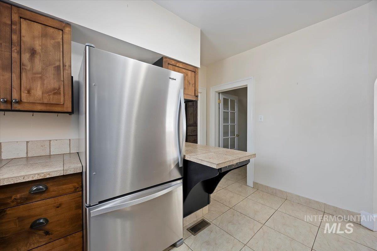Kitchen with freestanding refrigerator, light tile patterned floors, tile countertops, and brown cabinetry