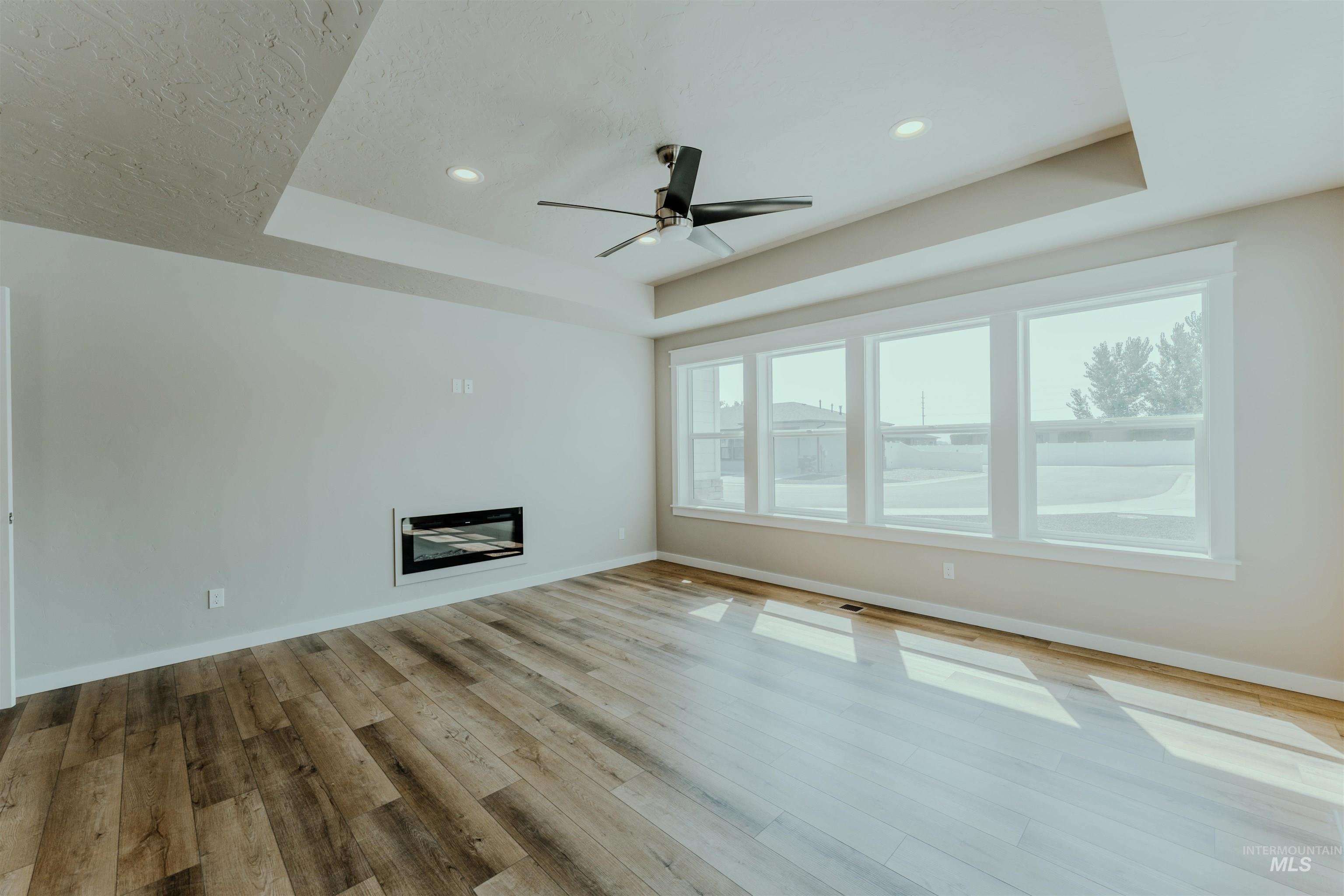 Unfurnished living room with a raised ceiling, a glass covered fireplace, a ceiling fan, light wood-style flooring, and recessed lighting