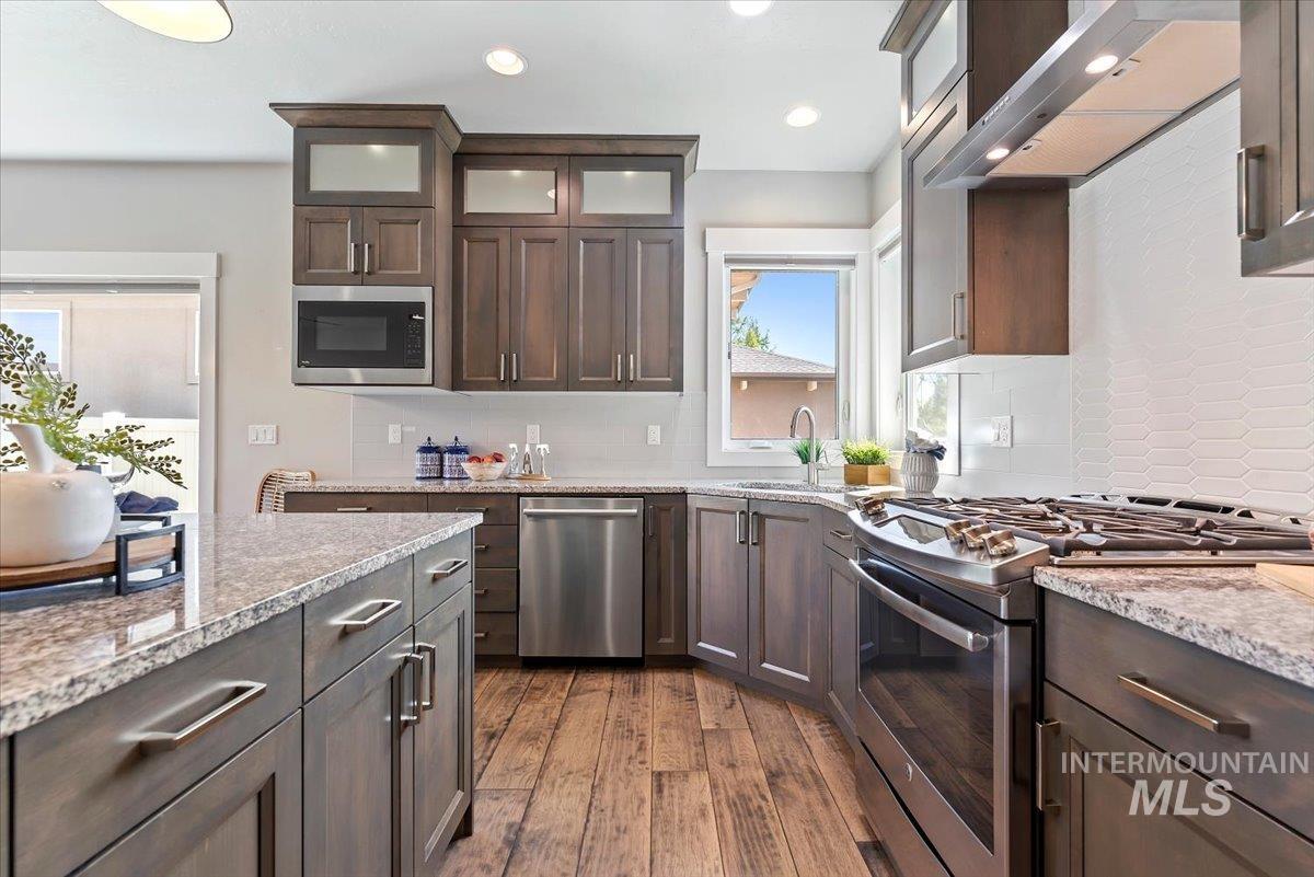 Kitchen with stainless steel appliances, dark brown cabinetry, wall chimney exhaust hood, light stone counters, and dark wood-style flooring