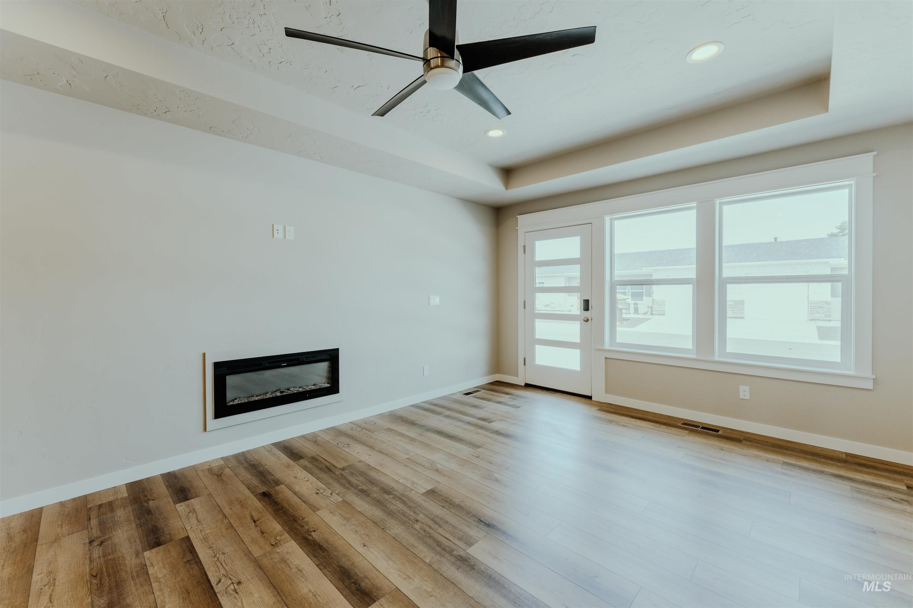Unfurnished living room featuring a raised ceiling, a glass covered fireplace, light wood-type flooring, ceiling fan, and recessed lighting