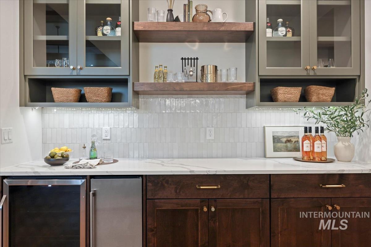 Indoor dry bar featuring beverage cooler, stainless steel refrigerator, tasteful backsplash, open shelves, and light stone counters