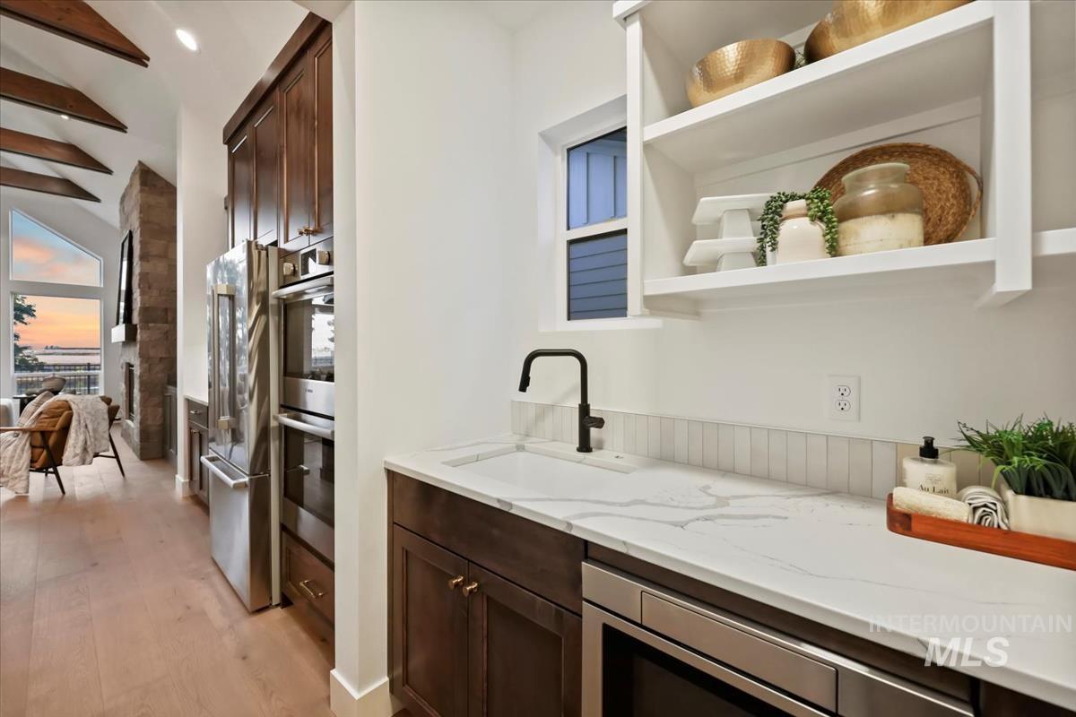 Bar area with dark brown cabinetry, open shelves, light stone counters, appliances with stainless steel finishes, and light wood-style flooring
