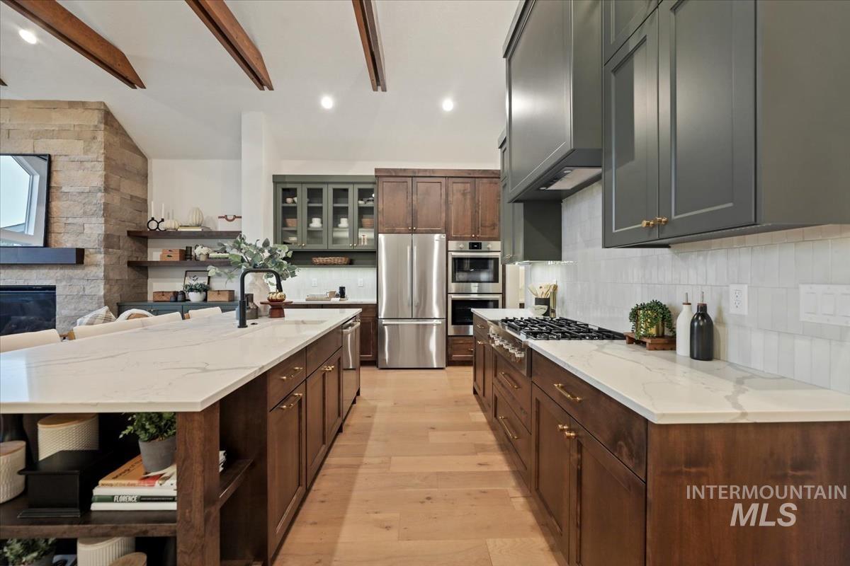 Kitchen with open shelves, light wood finished floors, light stone countertops, stainless steel appliances, and an island with sink