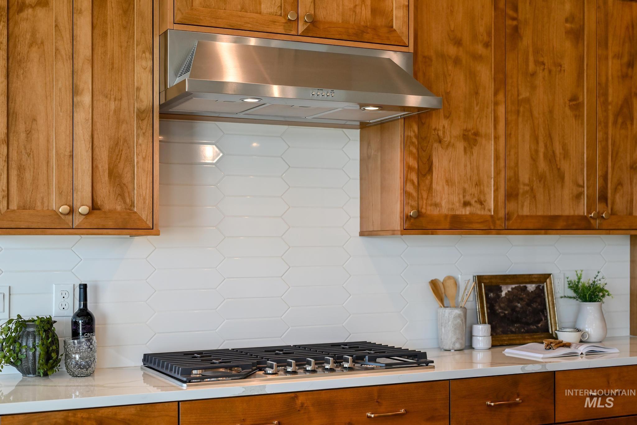 Kitchen featuring decorative backsplash, brown cabinetry, wall chimney range hood, stainless steel gas stovetop, and light stone counters