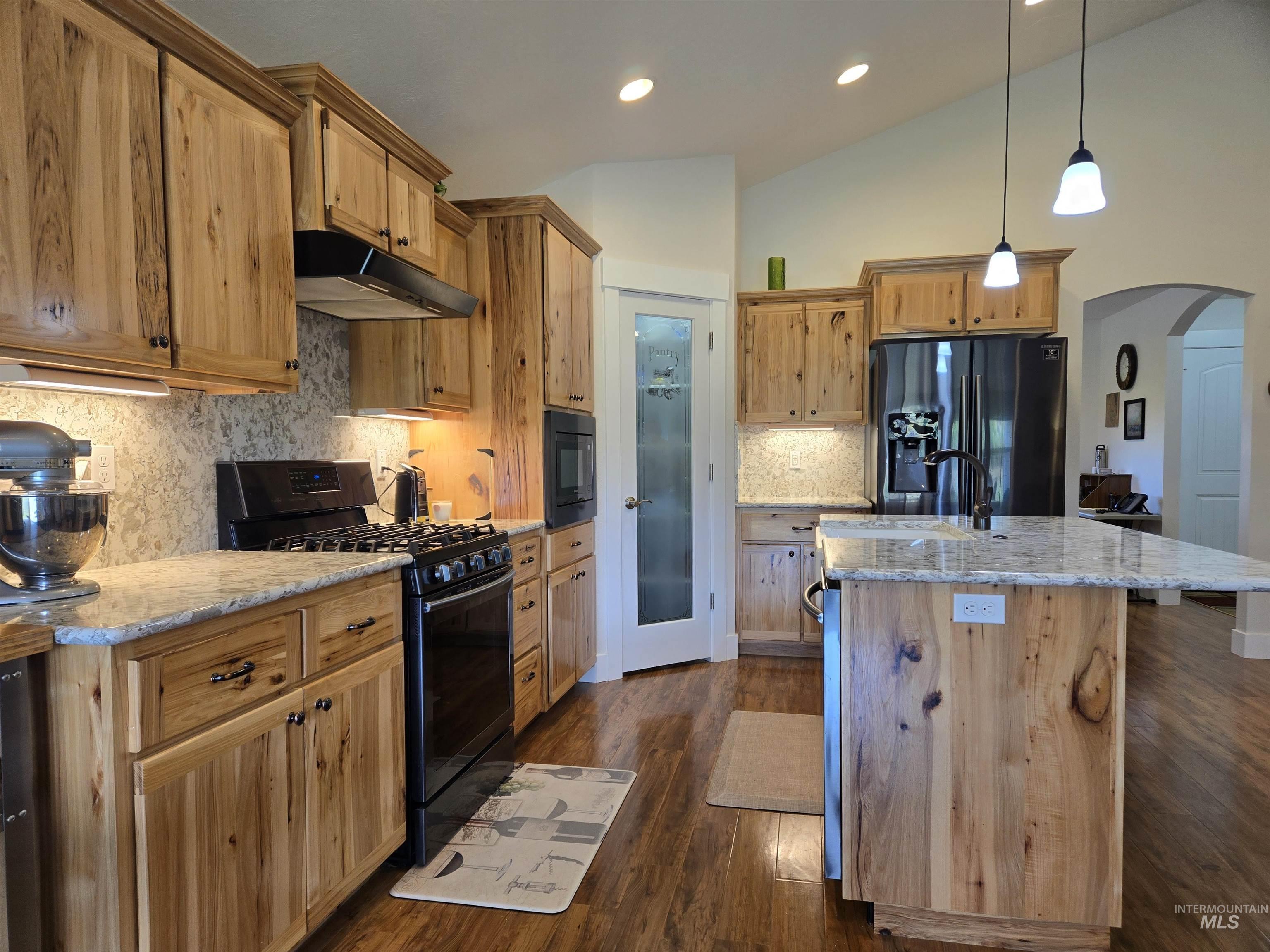 Kitchen with arched walkways, black appliances, backsplash, under cabinet range hood, and light stone countertops