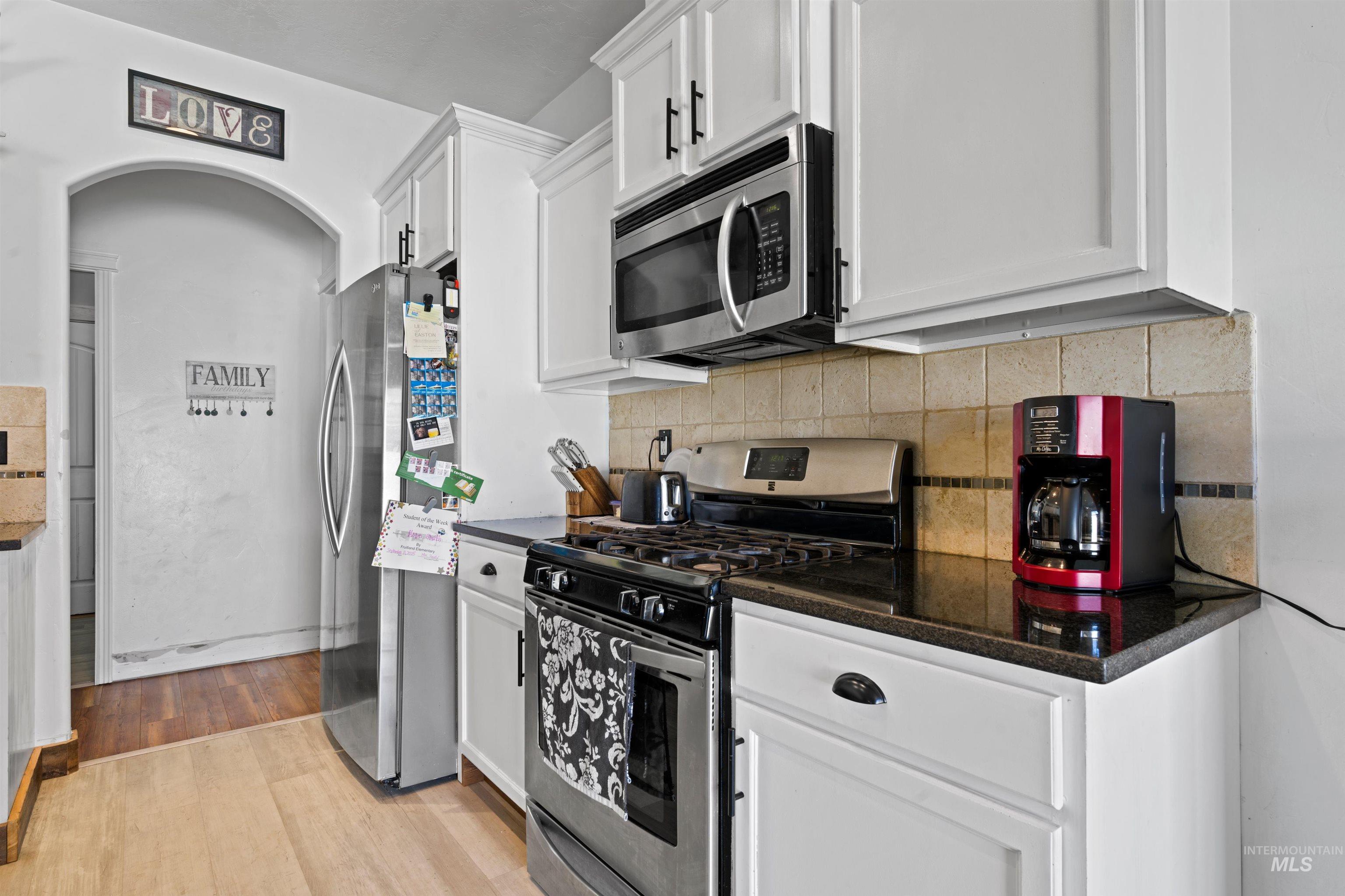 Kitchen with appliances with stainless steel finishes, white cabinets, backsplash, light wood finished floors, and dark stone countertops