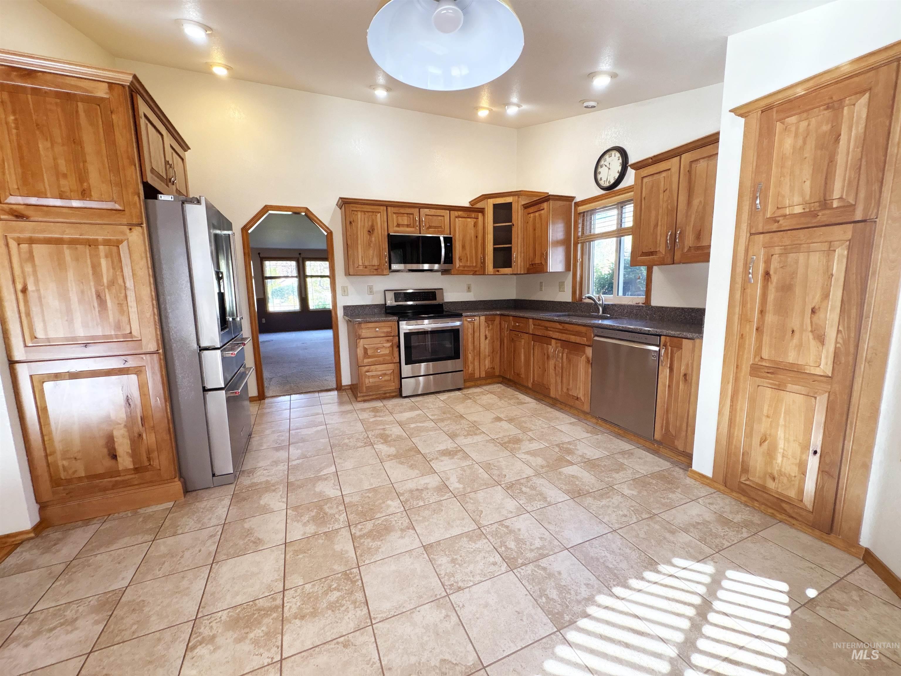 Kitchen with arched walkways, appliances with stainless steel finishes, brown cabinetry, glass insert cabinets, and light tile patterned flooring