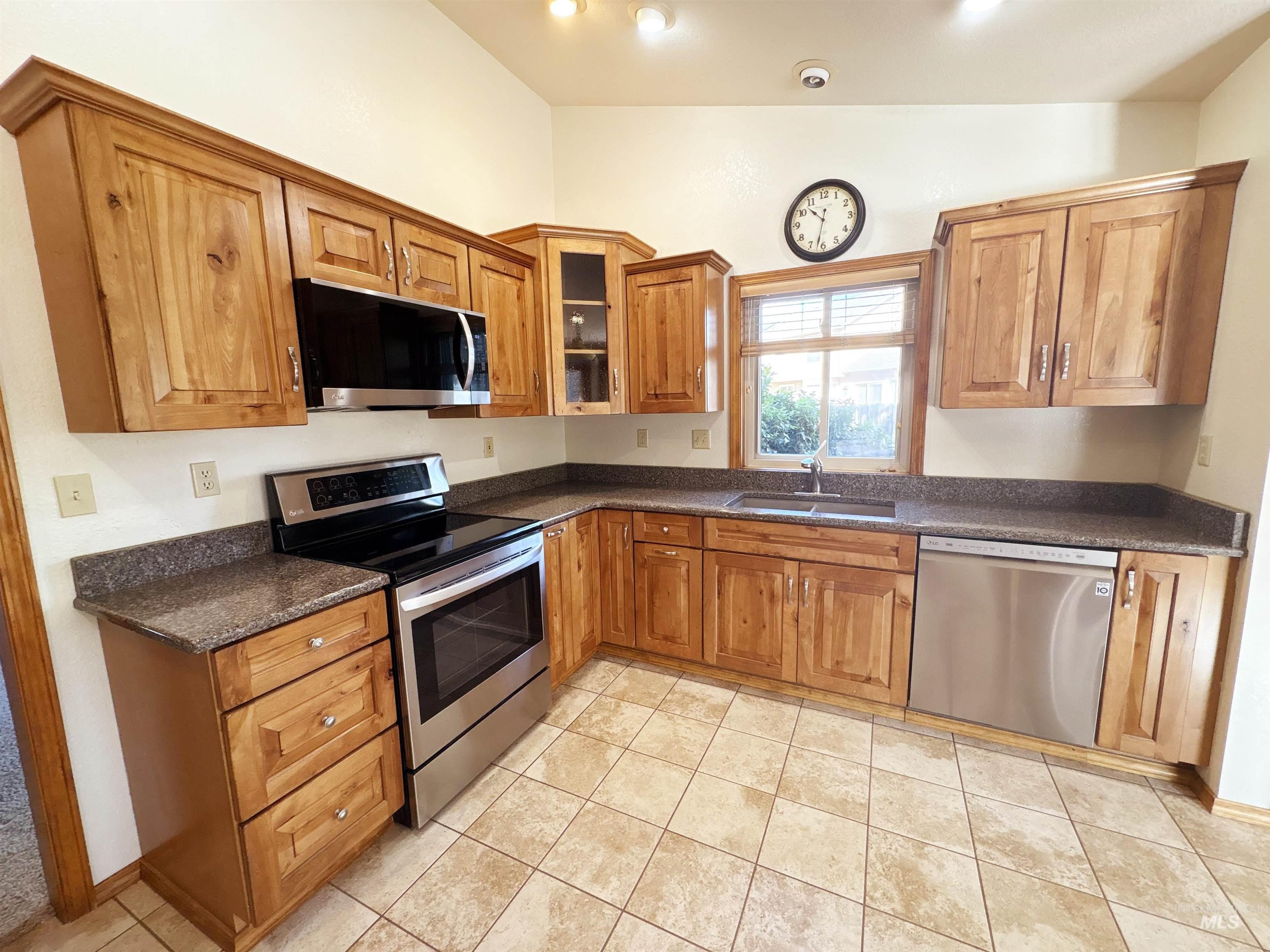 Kitchen with stainless steel appliances, light tile patterned floors, glass insert cabinets, brown cabinetry, and dark stone countertops