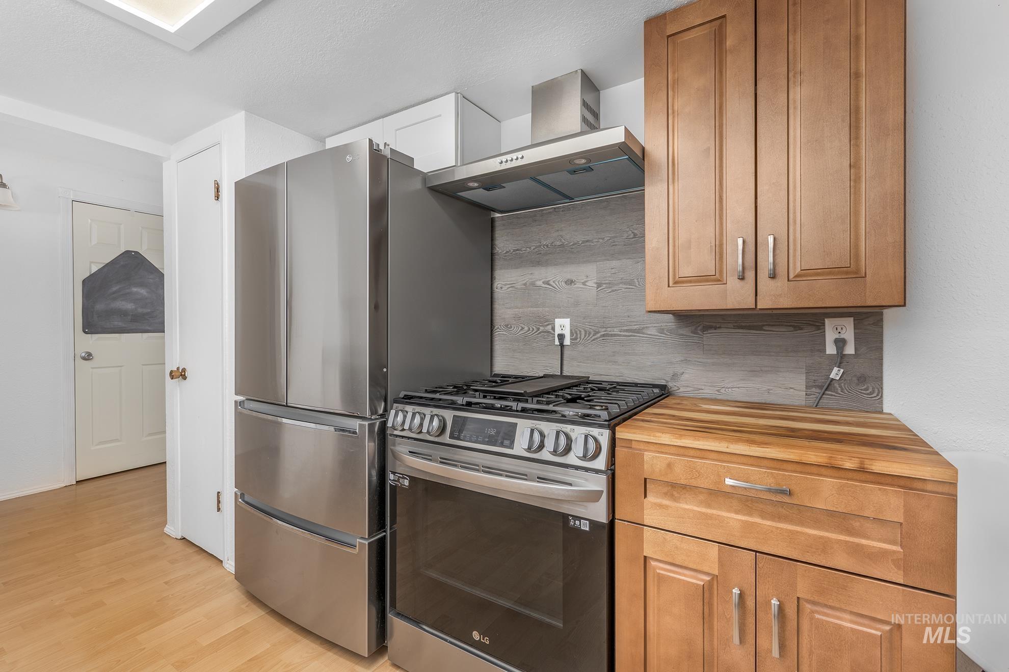 Kitchen featuring stainless steel range with gas stovetop, wall chimney exhaust hood, light wood-type flooring, backsplash, and brown cabinets