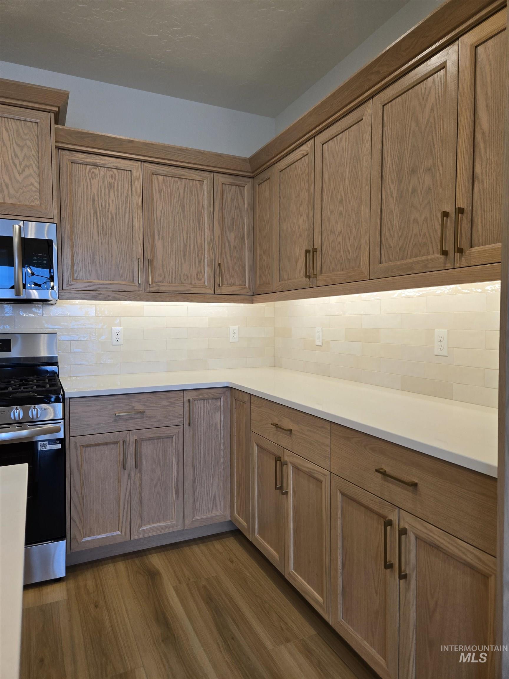Kitchen with appliances with stainless steel finishes, dark wood-type flooring, tasteful backsplash, brown cabinetry, and light stone countertops