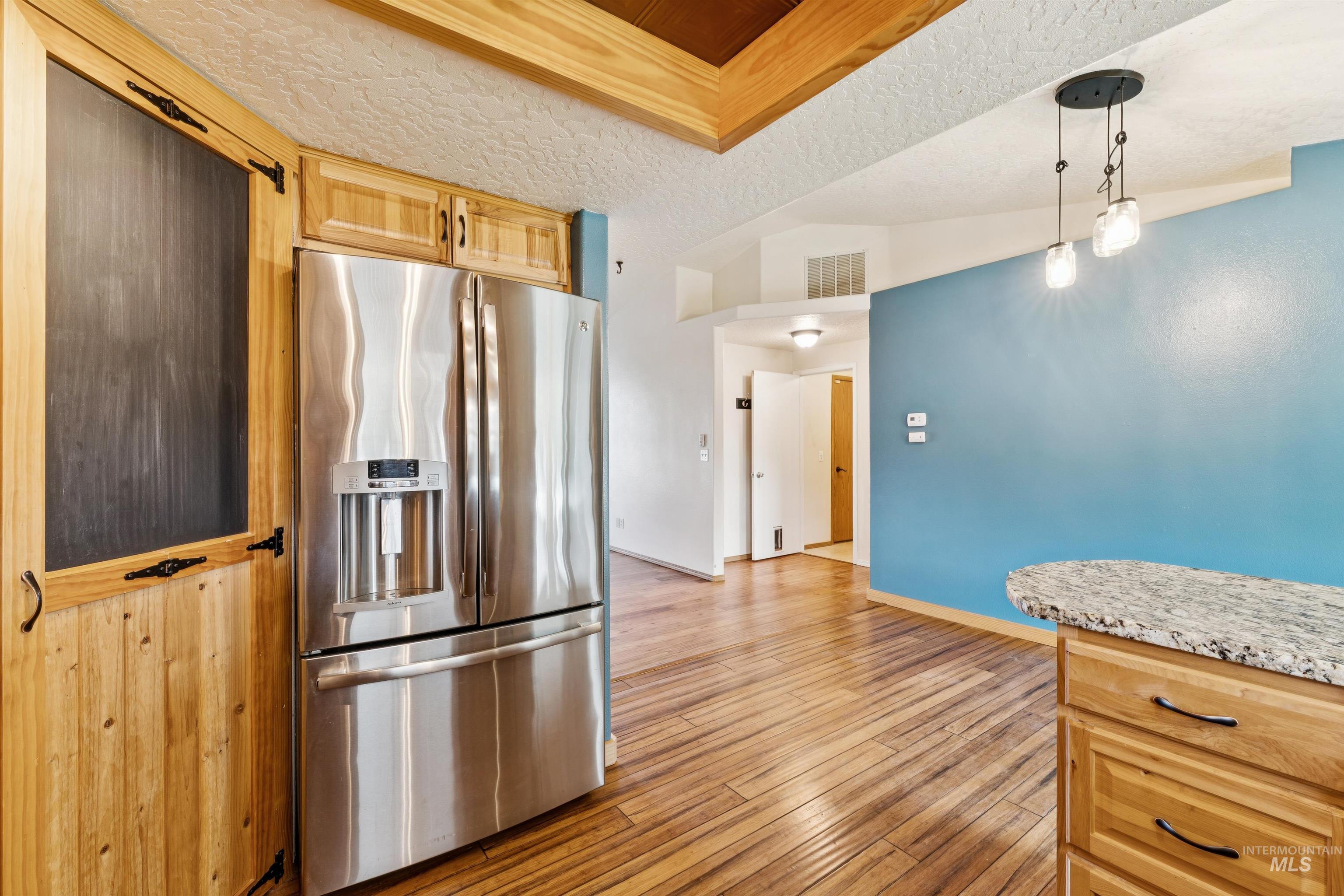 Kitchen featuring stainless steel fridge with ice dispenser, light brown cabinetry, hanging light fixtures, light wood-style flooring, and light stone counters