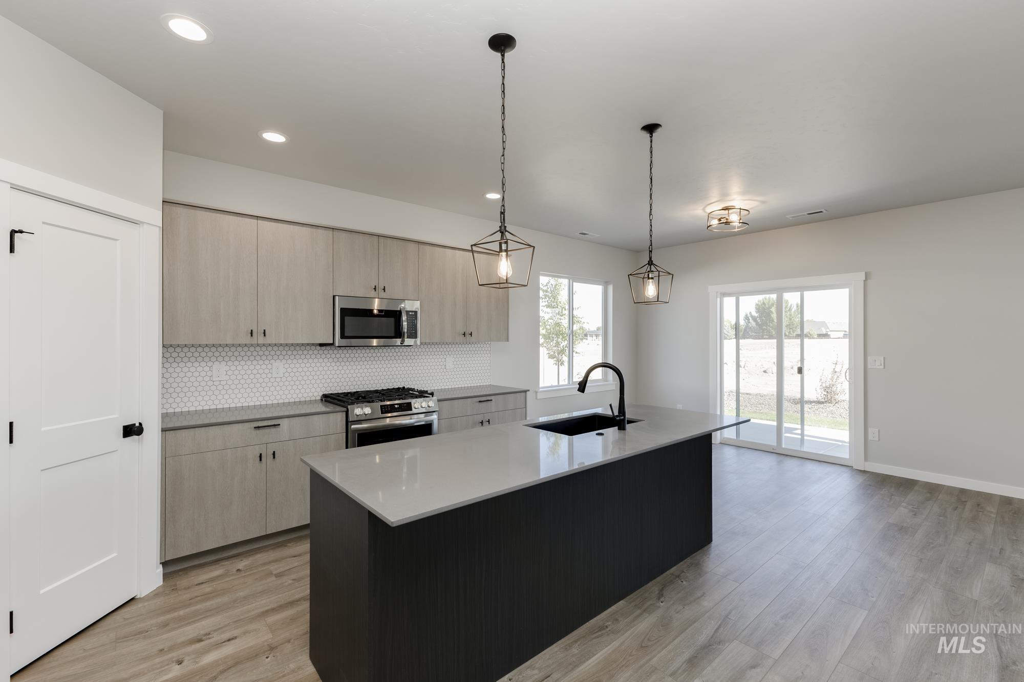 Kitchen featuring light brown cabinets, decorative light fixtures, a kitchen island with sink, tasteful backsplash, and light stone counters