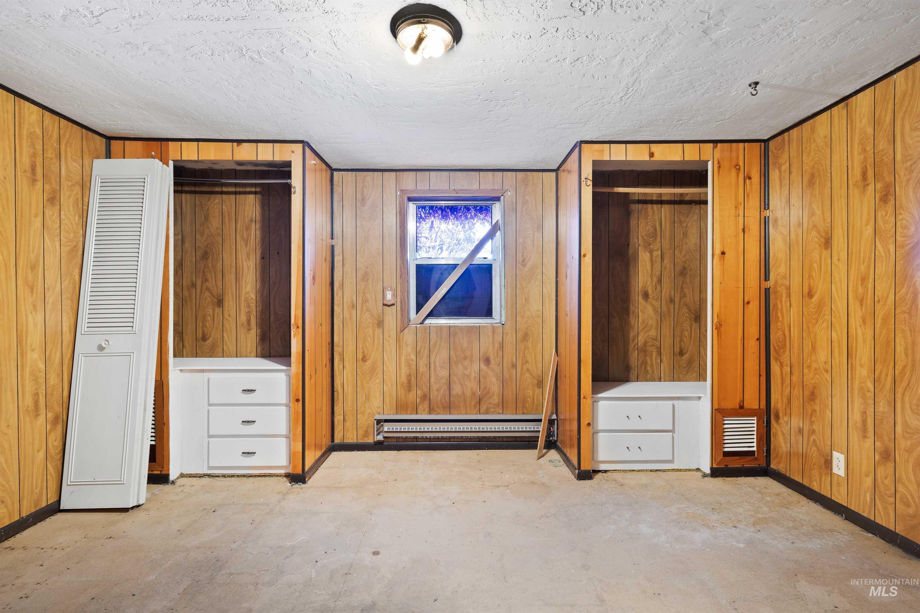 Unfurnished bedroom with wooden walls, a heating unit, multiple closets, unfinished concrete floors, and a baseboard radiator